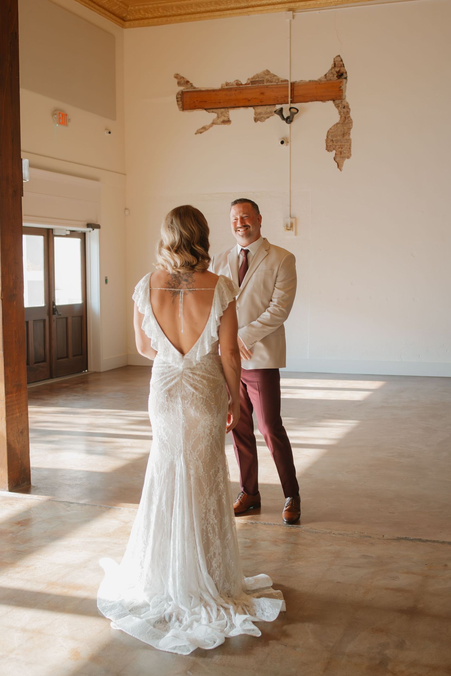 Groom smiles at bride in elegant backless lace gown during emotional first look in rustic venue.