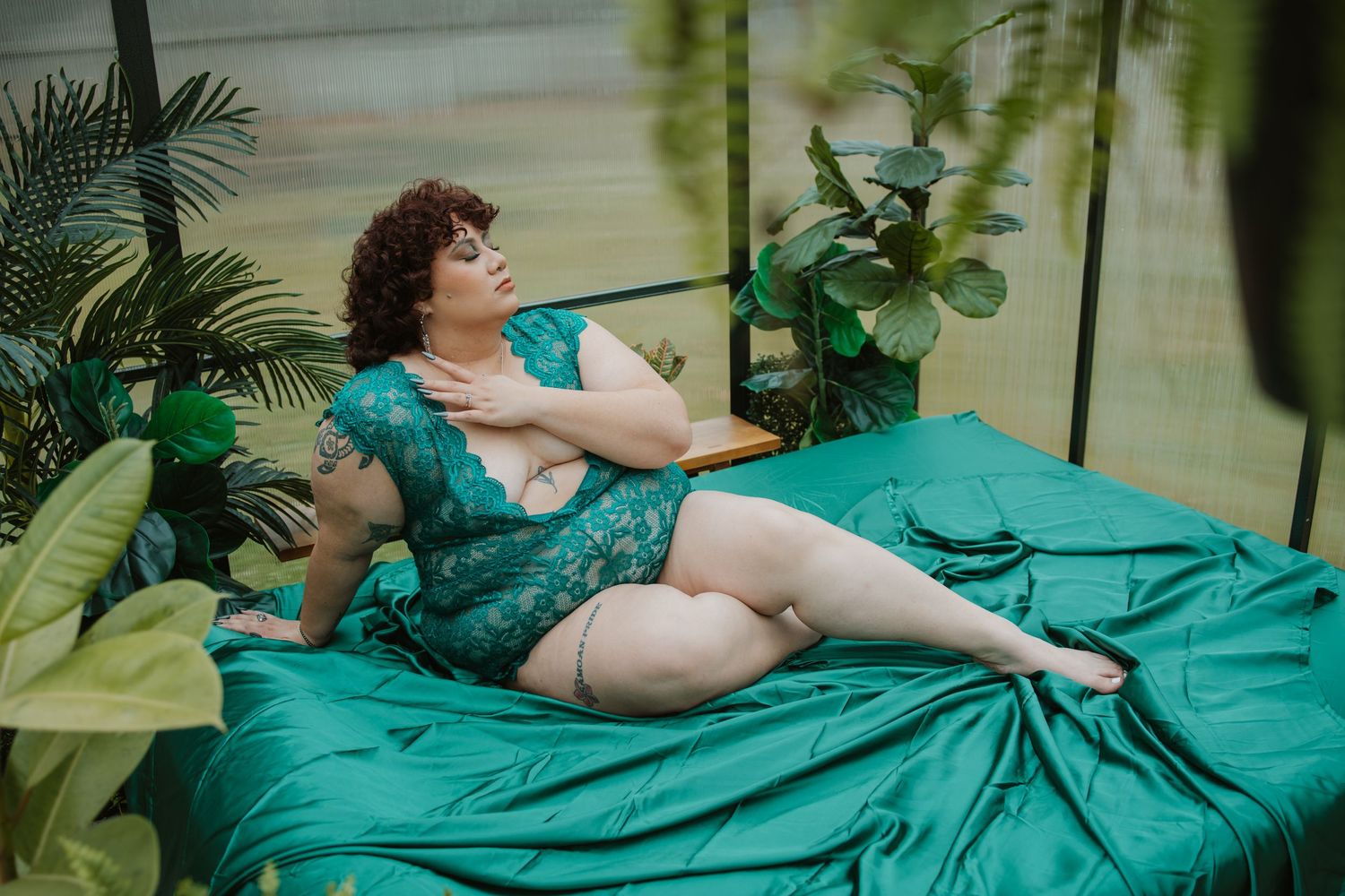 Plus-size woman in green lace lingerie relaxing on a teal bed surrounded by tropical plants.