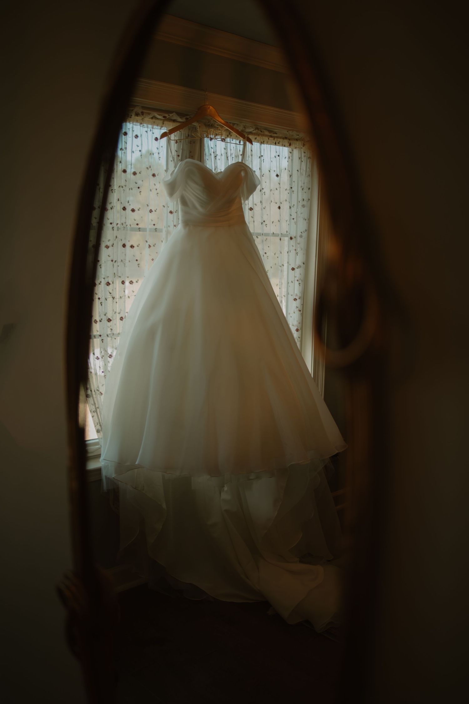 Elegant white ball gown wedding dress reflected in vintage oval mirror near a lace-curtained window.