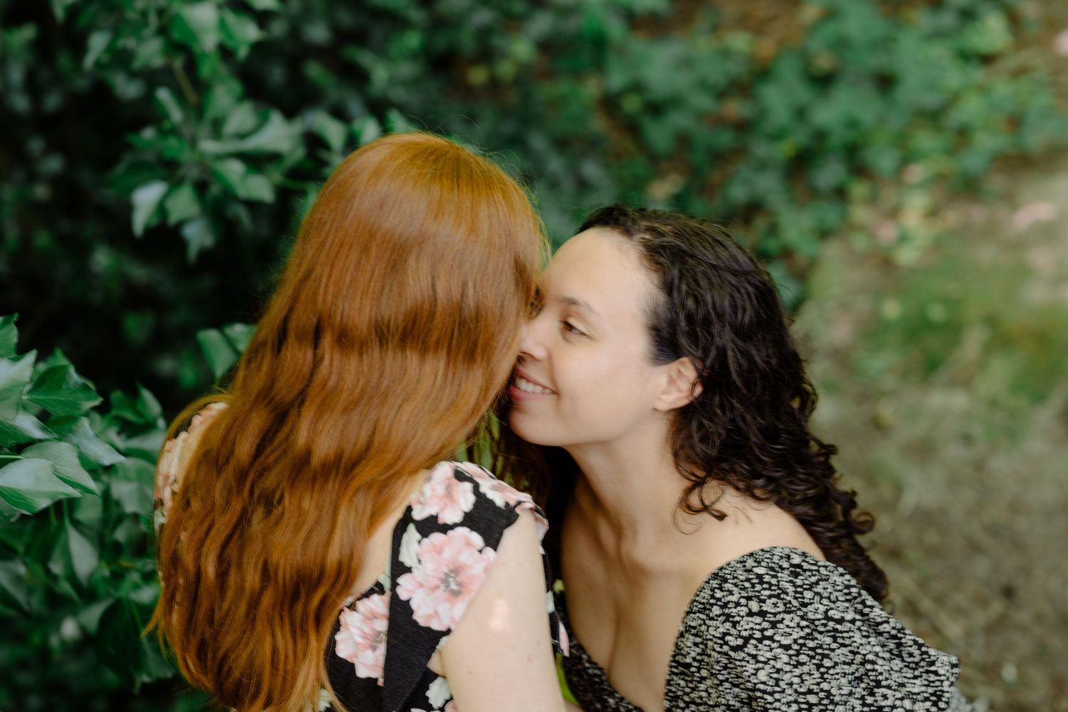 Two women sharing a tender moment outdoors, one with red hair and one with curly dark hair, surrounded by lush green foliage.