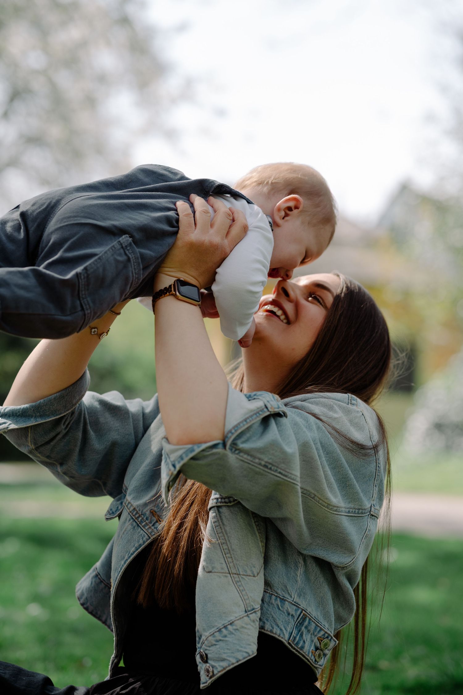 A young mother joyfully lifts her baby in the air while sitting in a green park on a sunny day. Mother and baby portrait in Eastbourne.