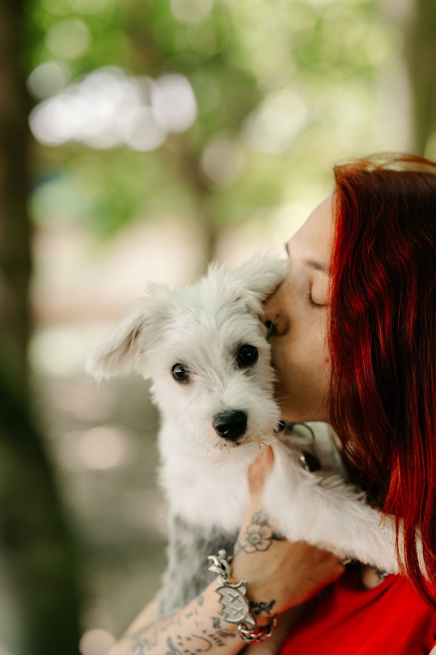 Red-haired woman with tattoos kissing a white fluffy dog outdoors in a green park setting.