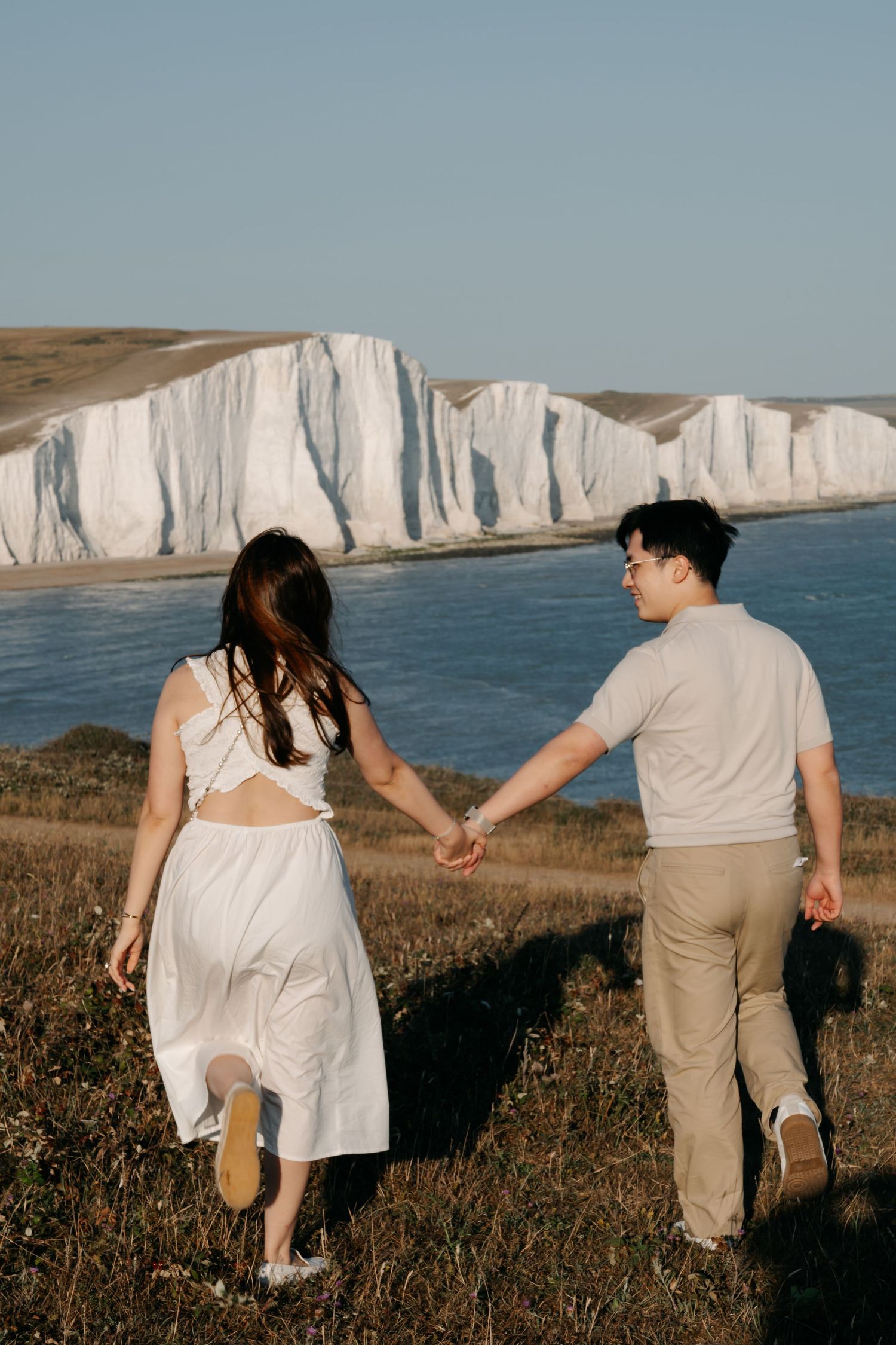 Couple holding hands walking toward white chalk cliffs by the sea on a sunny day.