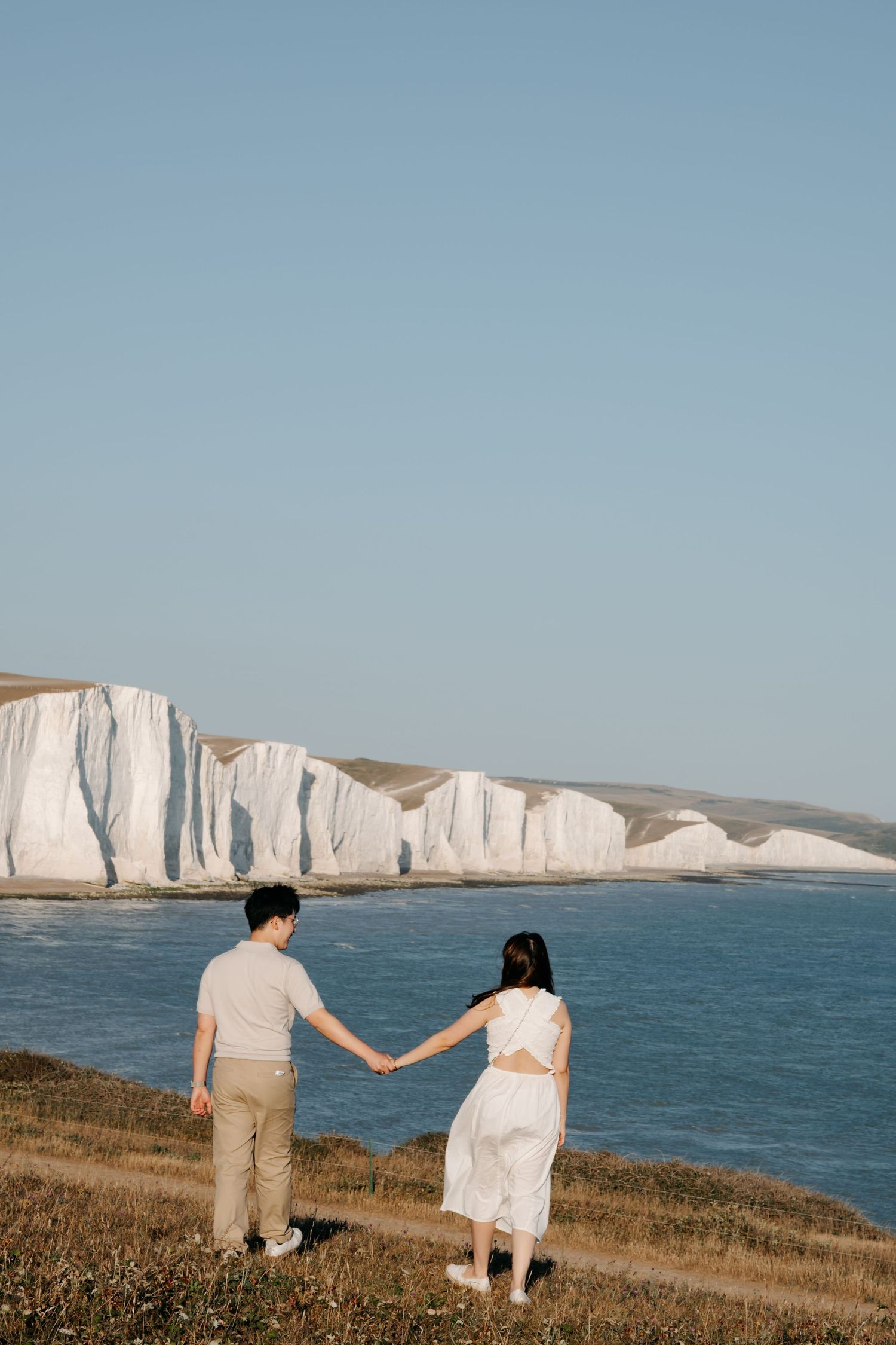 Couple holding hands facing white chalk cliffs by the sea on a clear sunny day.