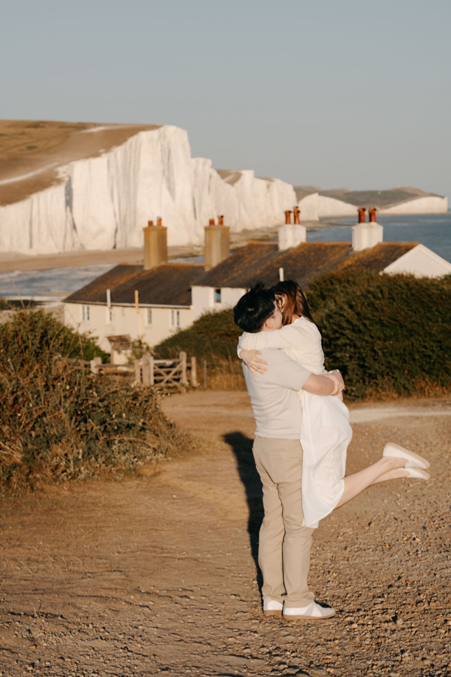 Couple embracing on a coastal path with white chalk cliffs and cottages in the background at golden hour.