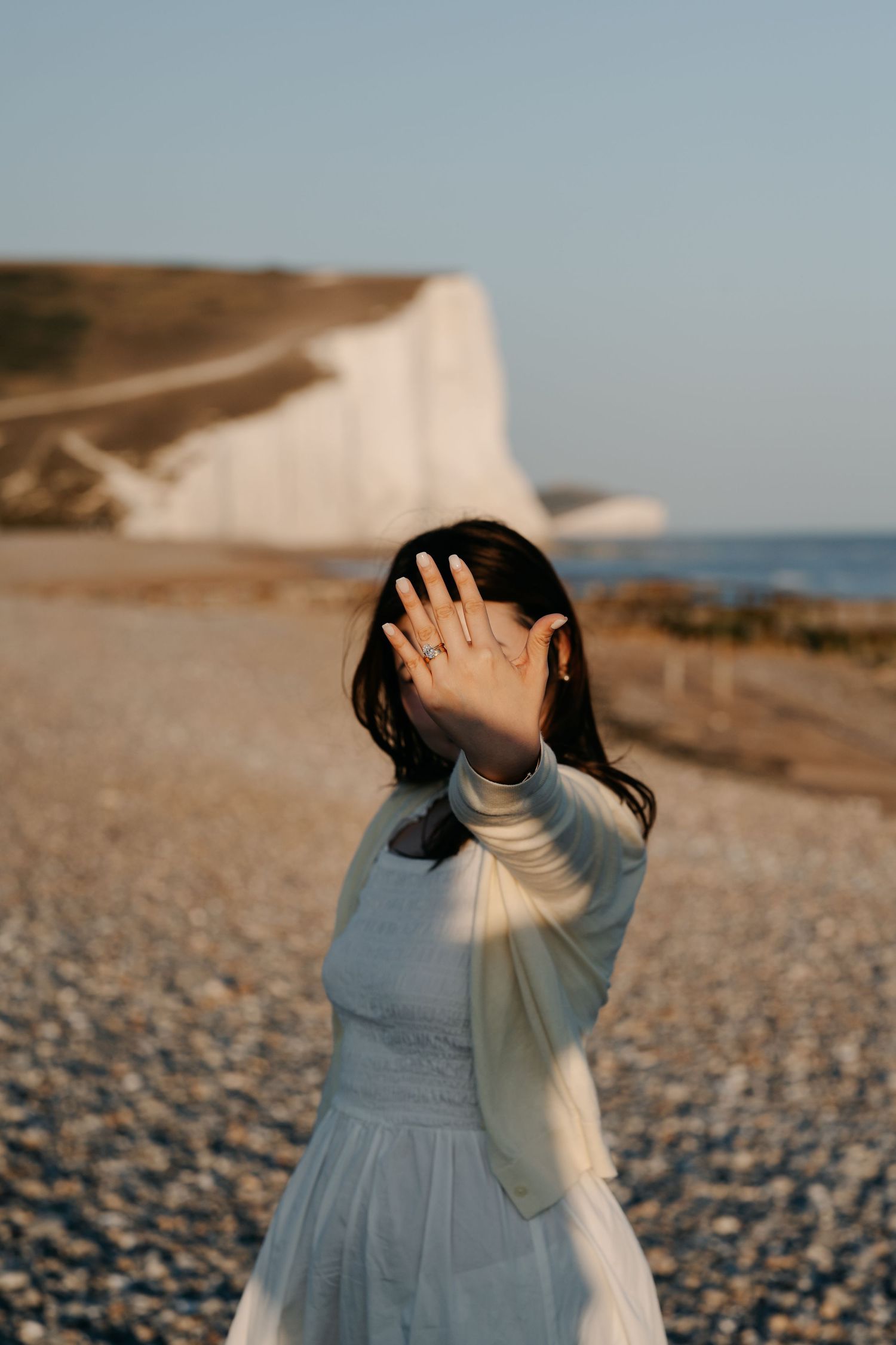 Woman in white dress on pebble beach showing engagement ring, with white cliffs and blue sky in background.