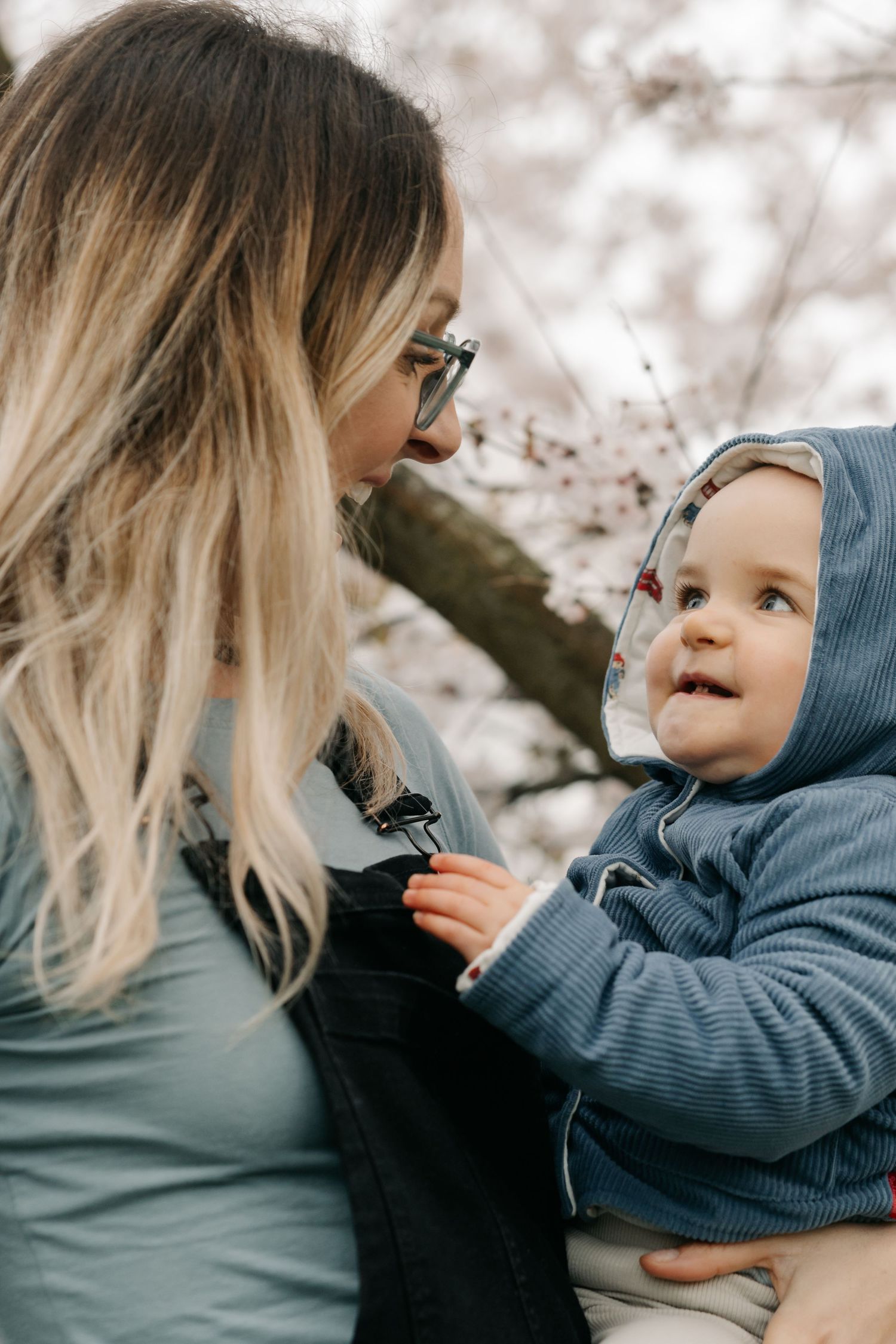 A mother holds her smiling baby in near cherry blossom trees in spring during a mother and baby portrait session in Eastbourne.