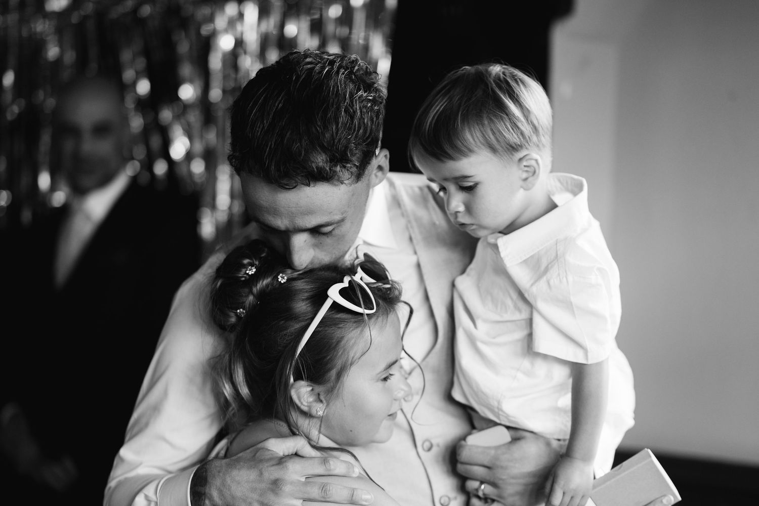 Black and white photo of a man kissing a young girl while holding a toddler boy at a wedding celebration.