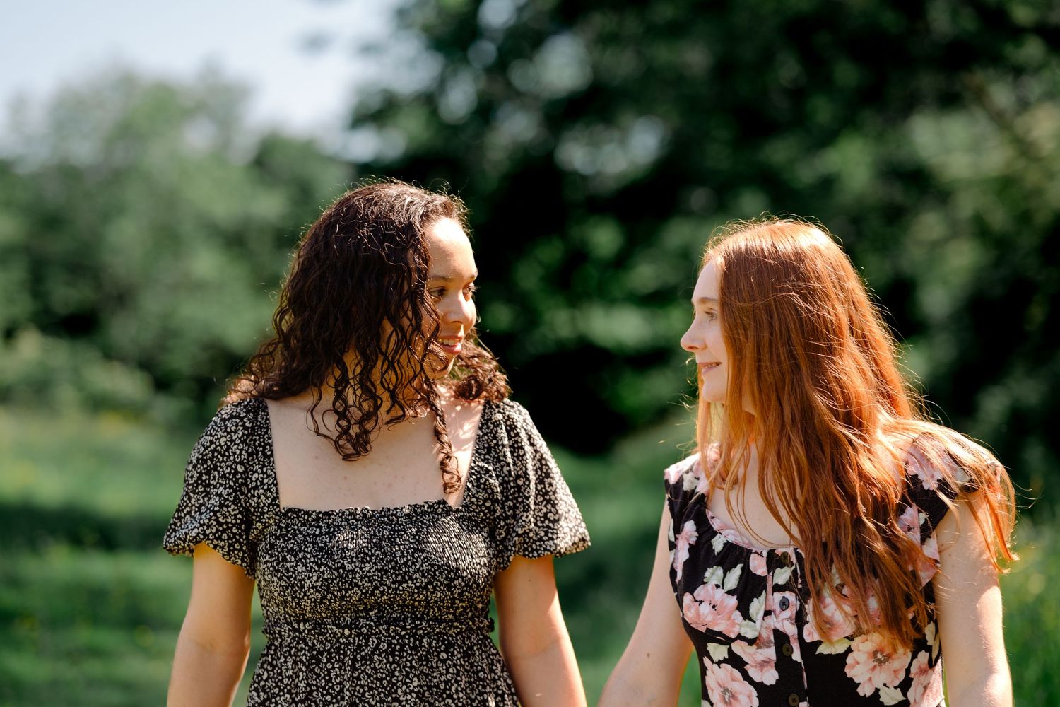 Two young women with curly hair walking outdoors, smiling at each other in a lush green park setting. Couple lgbt photography eastbourne