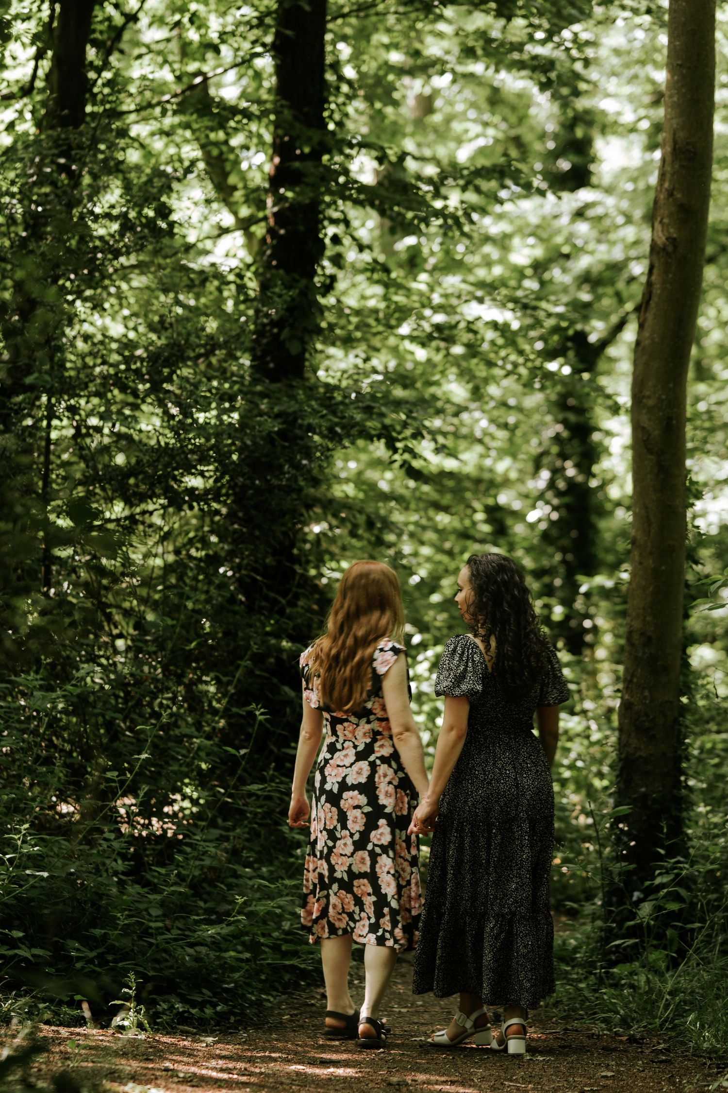 Two women in floral dresses walk hand in hand along a sunlit forest path surrounded by lush green trees in LGBT couple photoshoot in Sussex