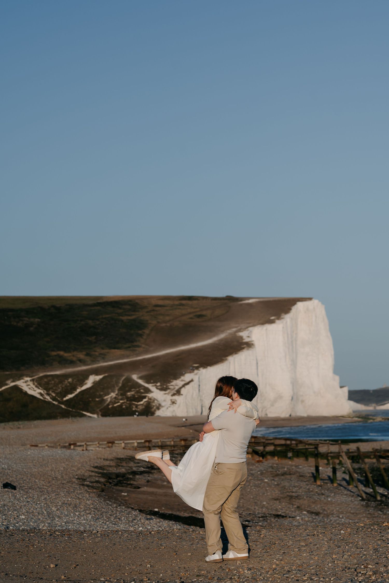 Couple embracing on a pebble beach with iconic white chalk cliffs in the background under a clear blue sky.