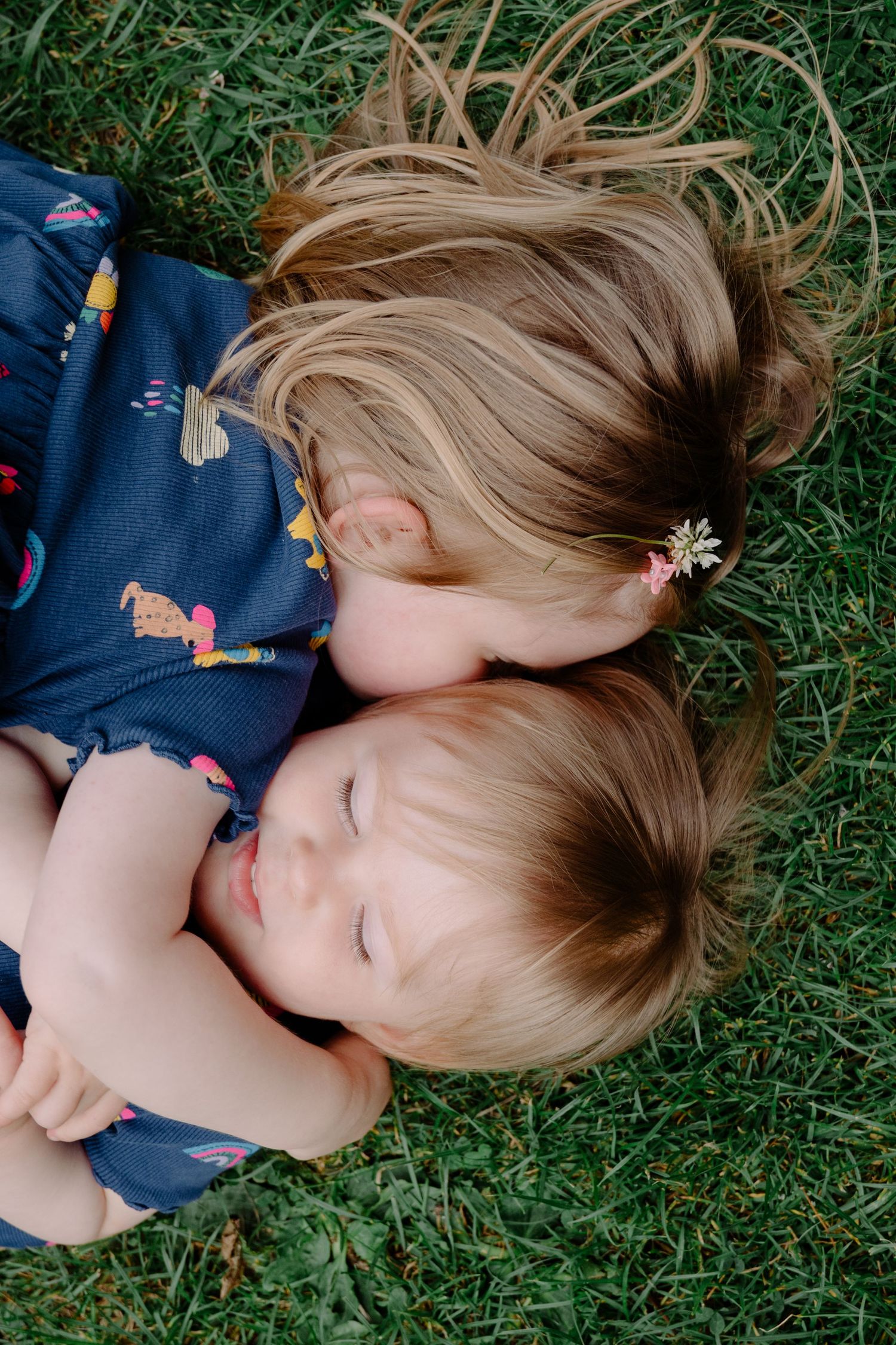 Two young blonde girls lying on green grass, one kissing the other's cheek in a sweet sibling moment.