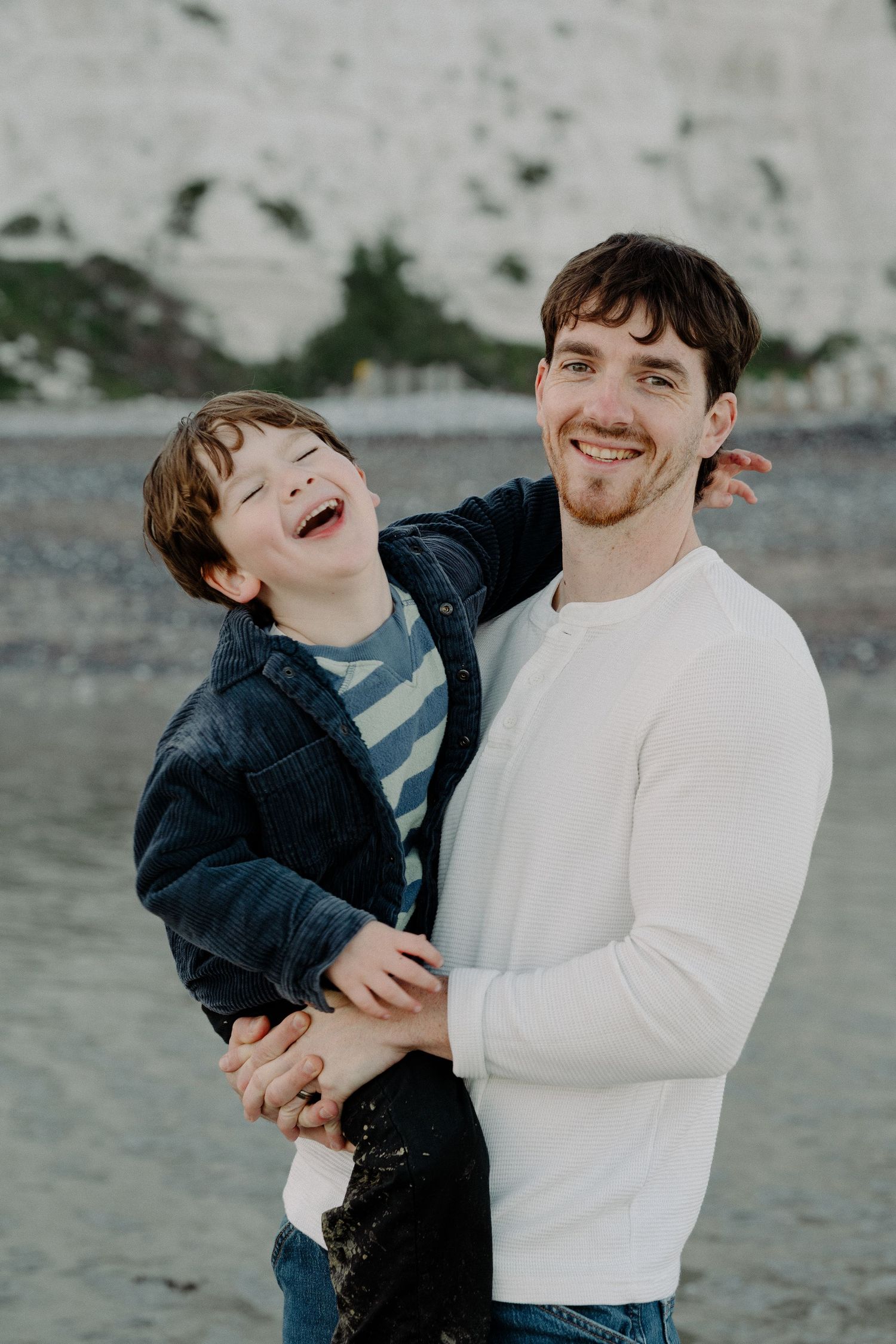 A smiling man holds a laughing young boy on a beach with white cliffs in the background. Family photoshoot eastbourne beach.