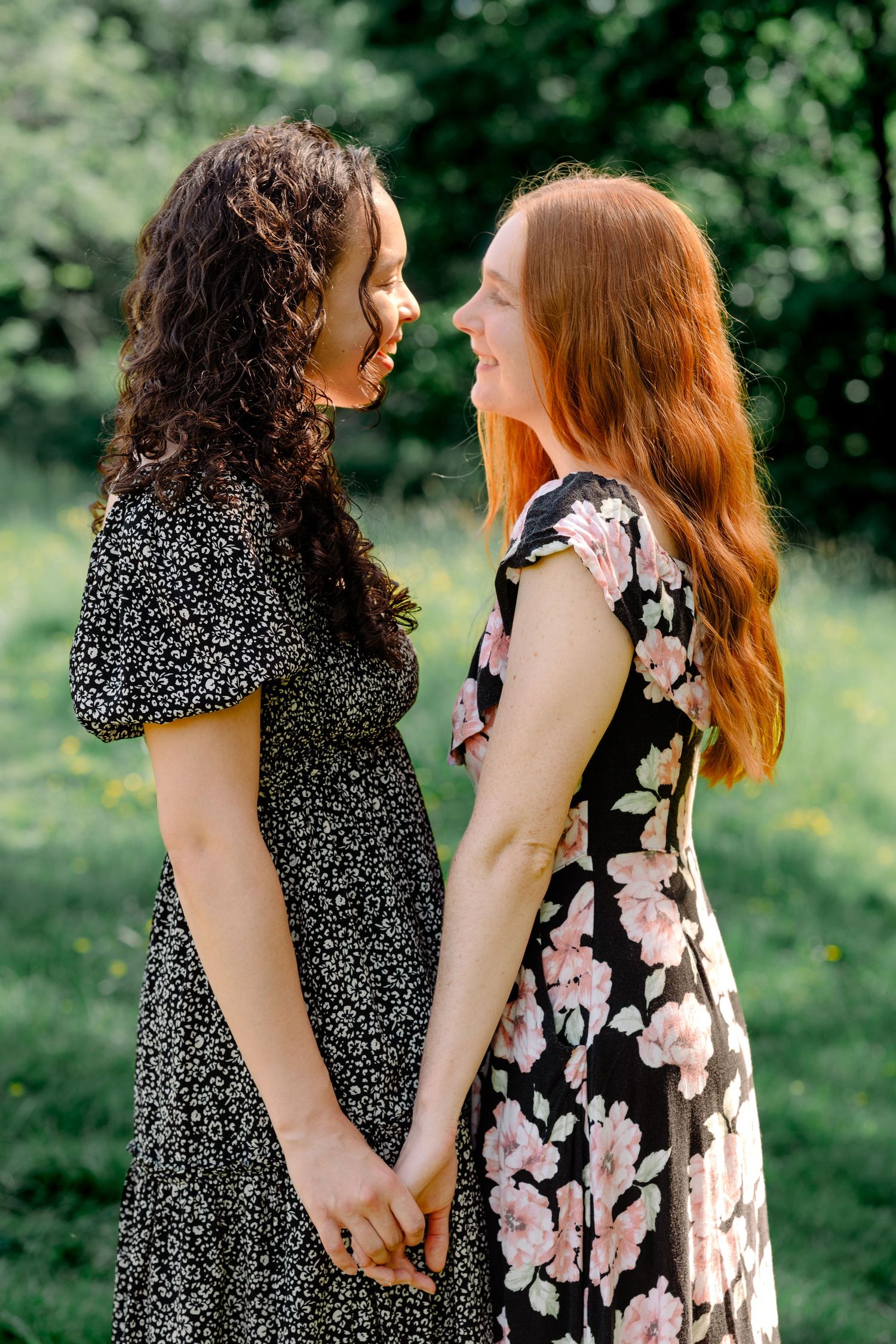 Two women in floral dresses hold hands and smile at each other in a sunny garden during a couple photography portrait session in Sussex.