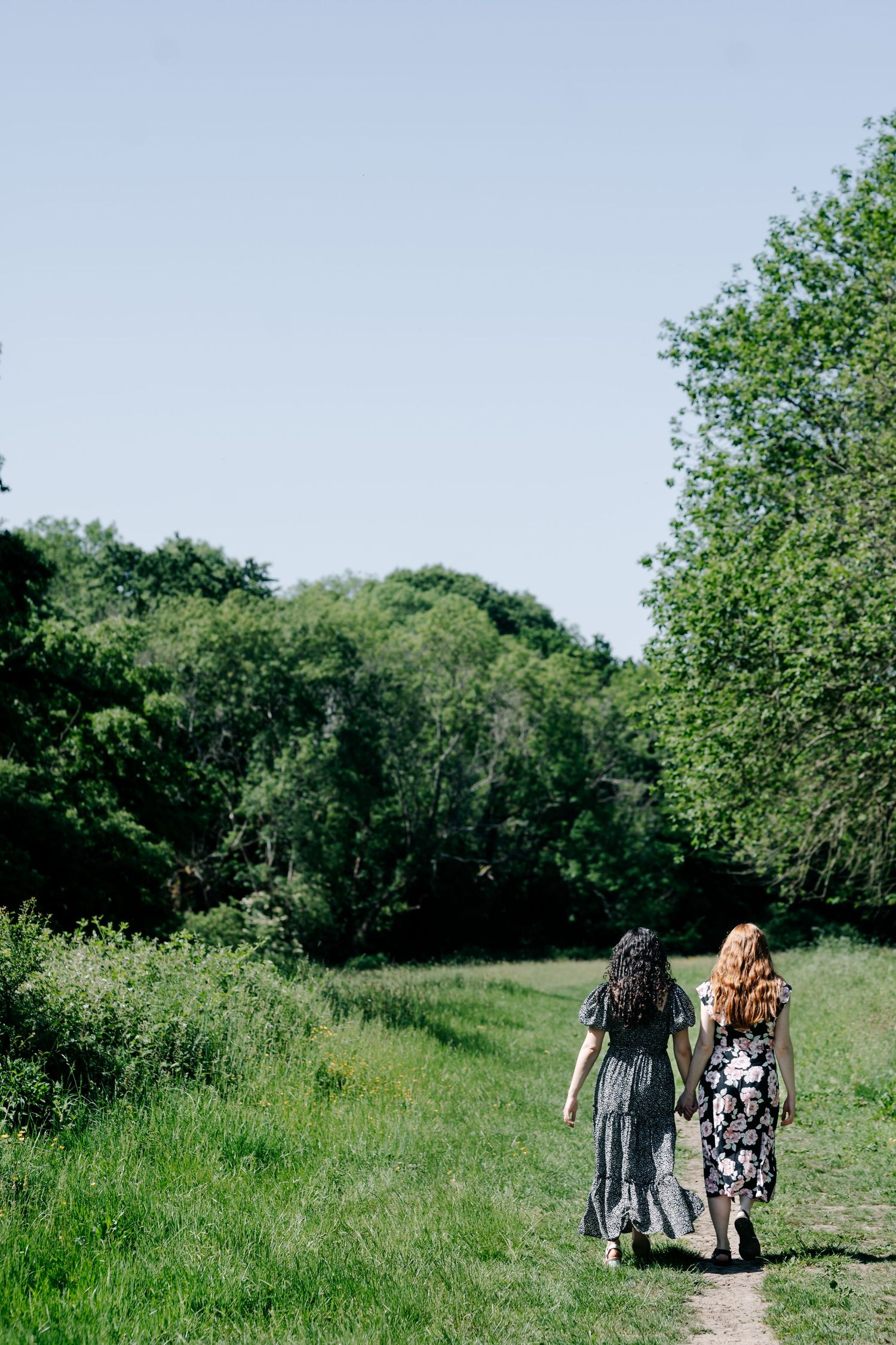 Two women in floral dresses walk hand in hand along a sunny green nature path surrounded by lush trees. Couple LGBT photography sussex