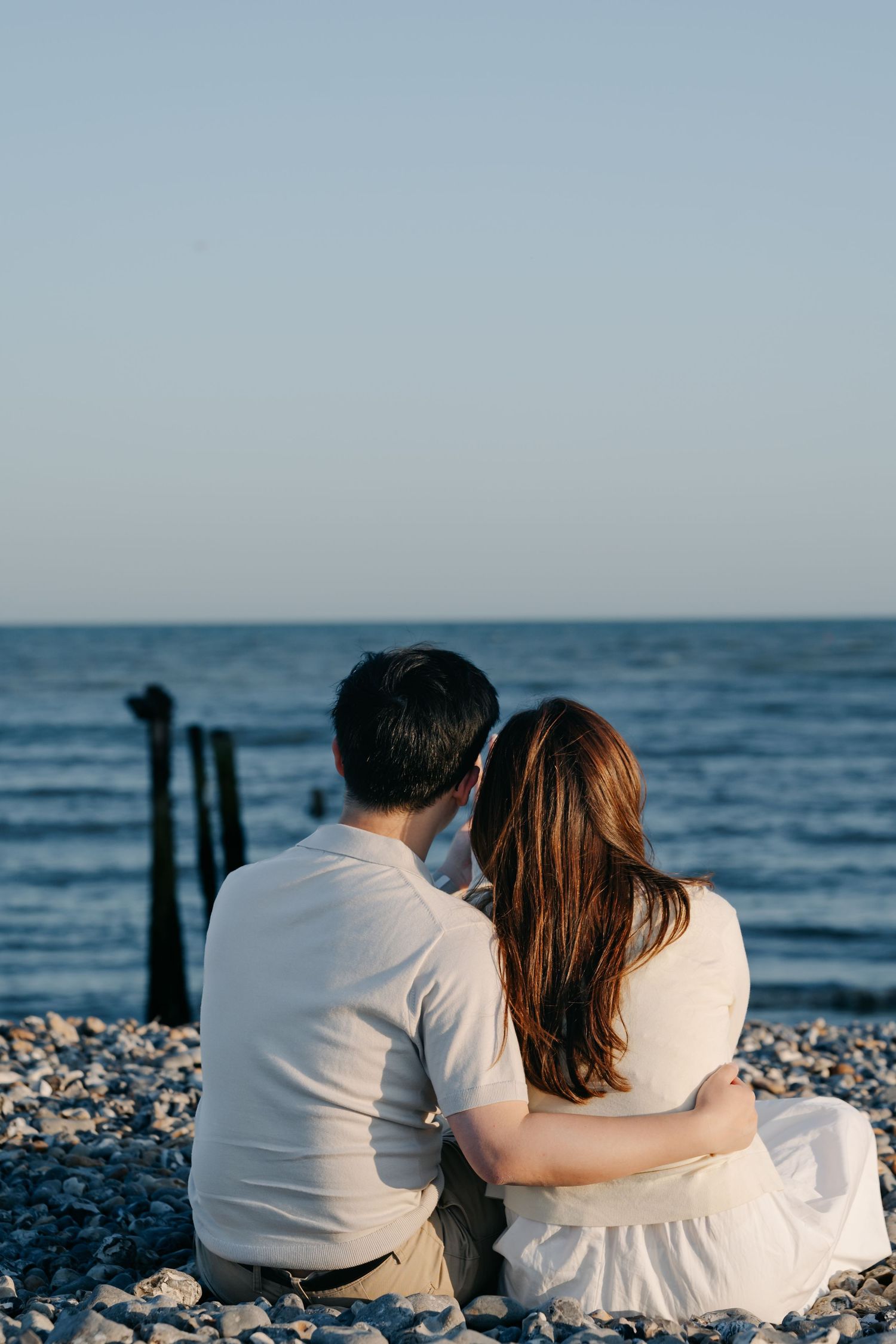 Couple sitting together on a rocky beach, embracing while gazing at the calm blue ocean horizon.