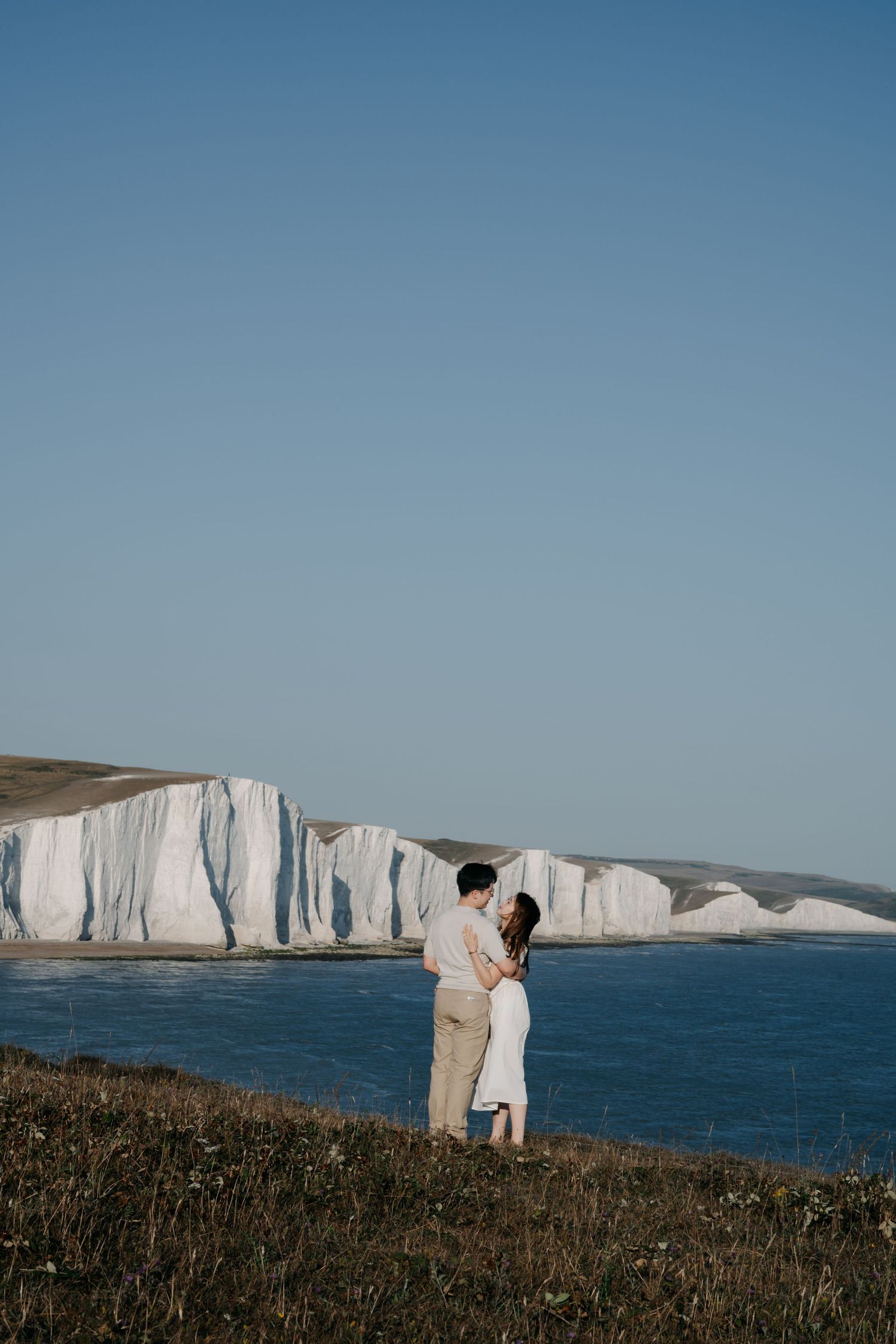 Couple embracing on coastal cliffs overlooking the white chalk Seven Sisters cliffs and blue sea in East Sussex, England.