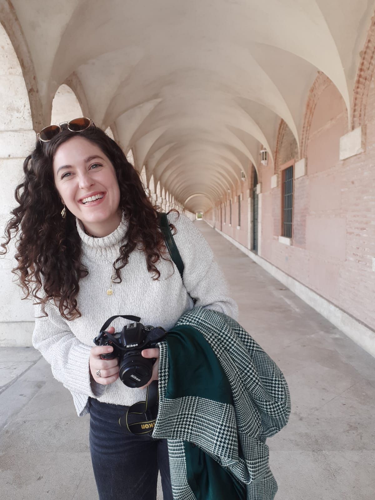 Smiling woman with curly hair holds a camera in an arched stone corridor, wearing a white sweater and carrying a plaid scarf.