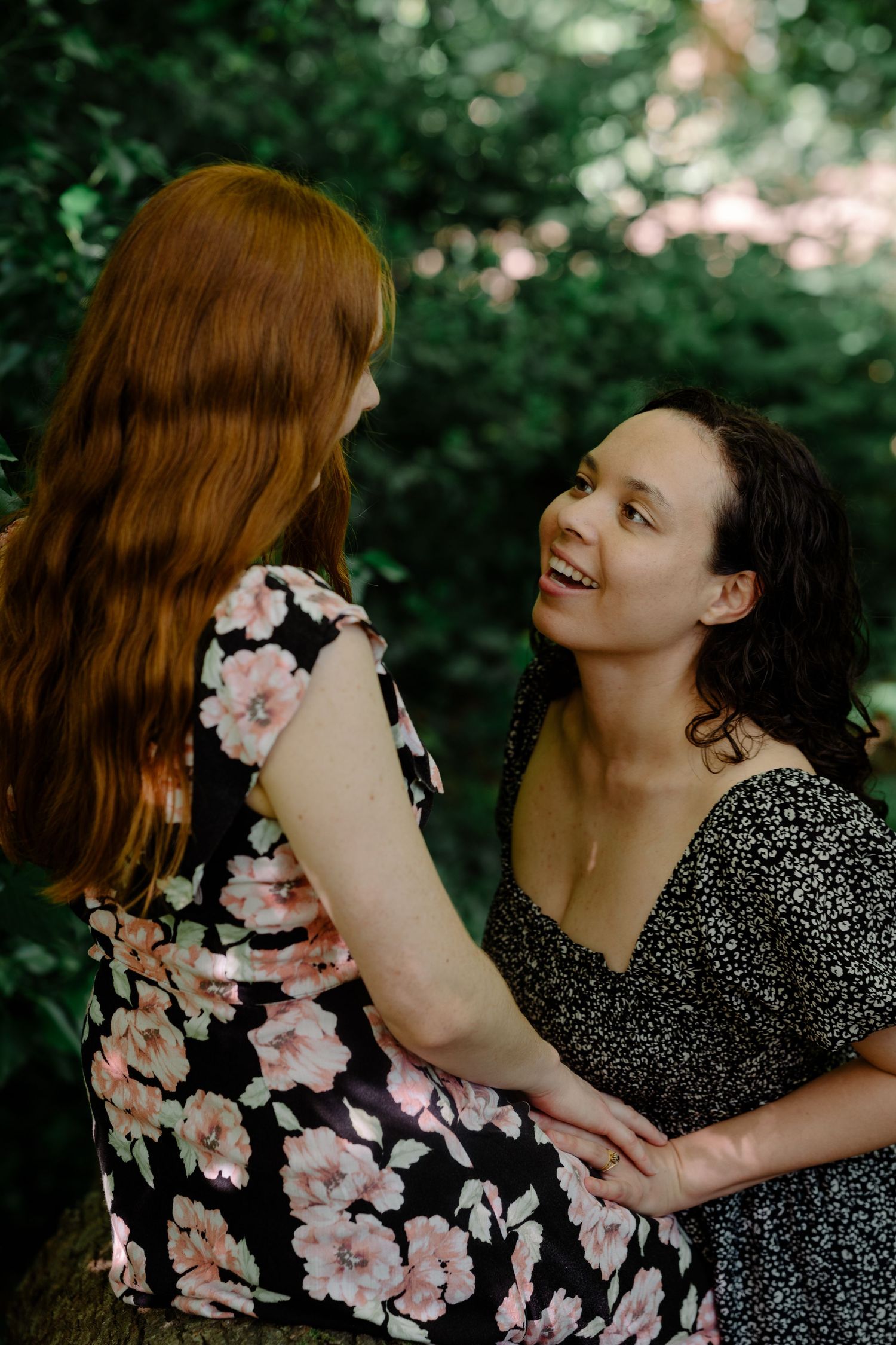 Two women in floral dresses share a joyful moment together surrounded by lush greenery during and LGBT couple portrait session in Sussex.