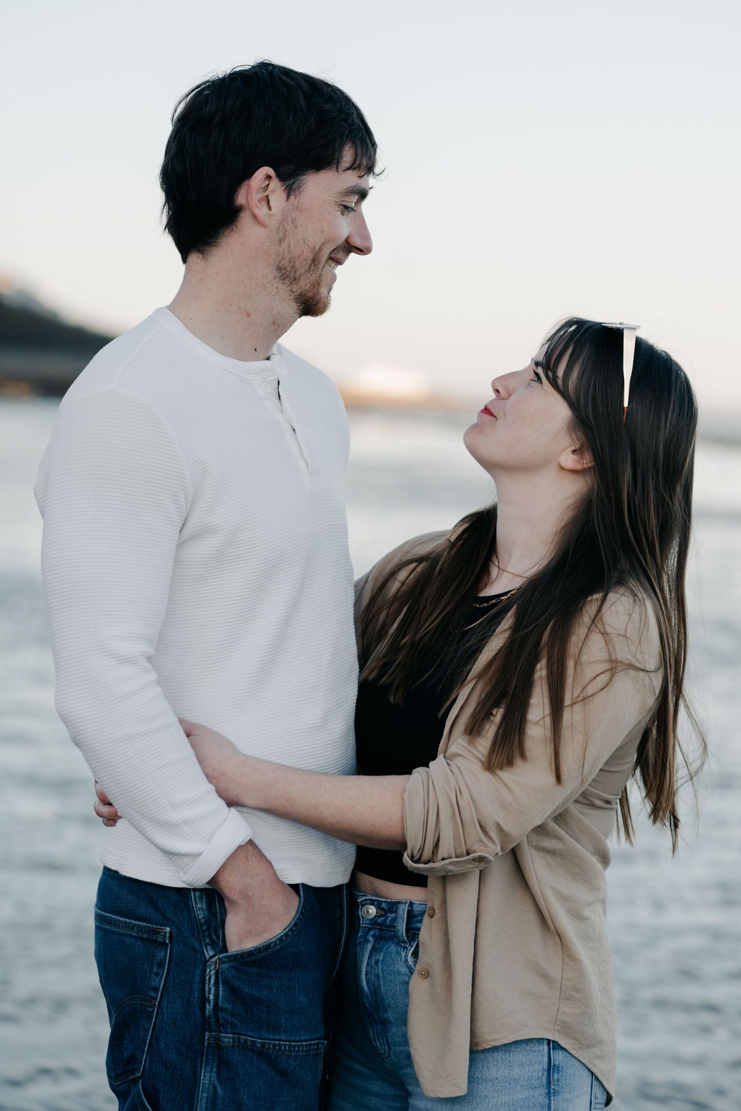 A happy couple embracing and smiling at each other on a beach at sunset, dressed casually in jeans and tops. Couple photoshoot sussex