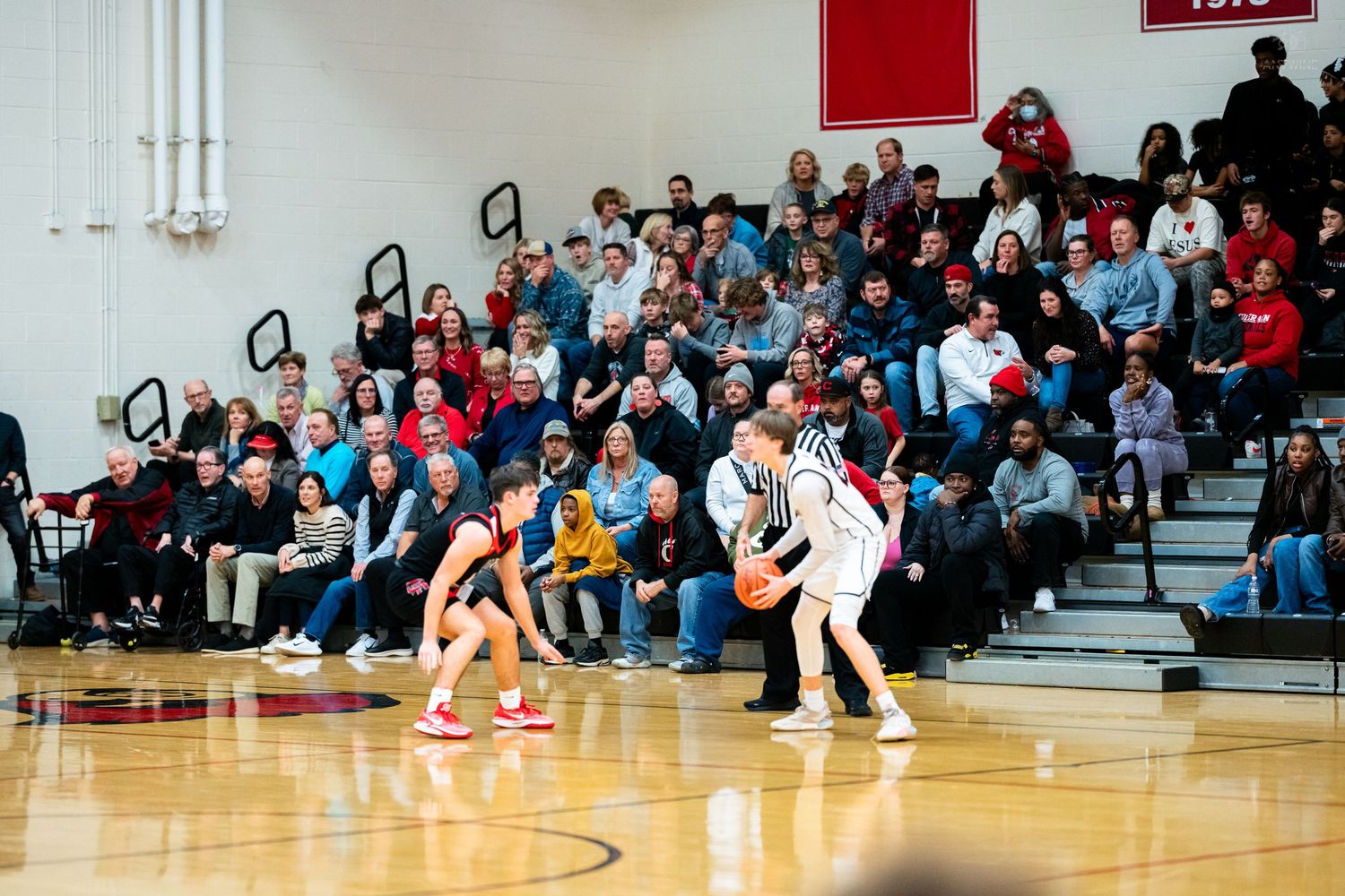 Two high school basketball players face off near sideline with large crowd of spectators in bleachers.
