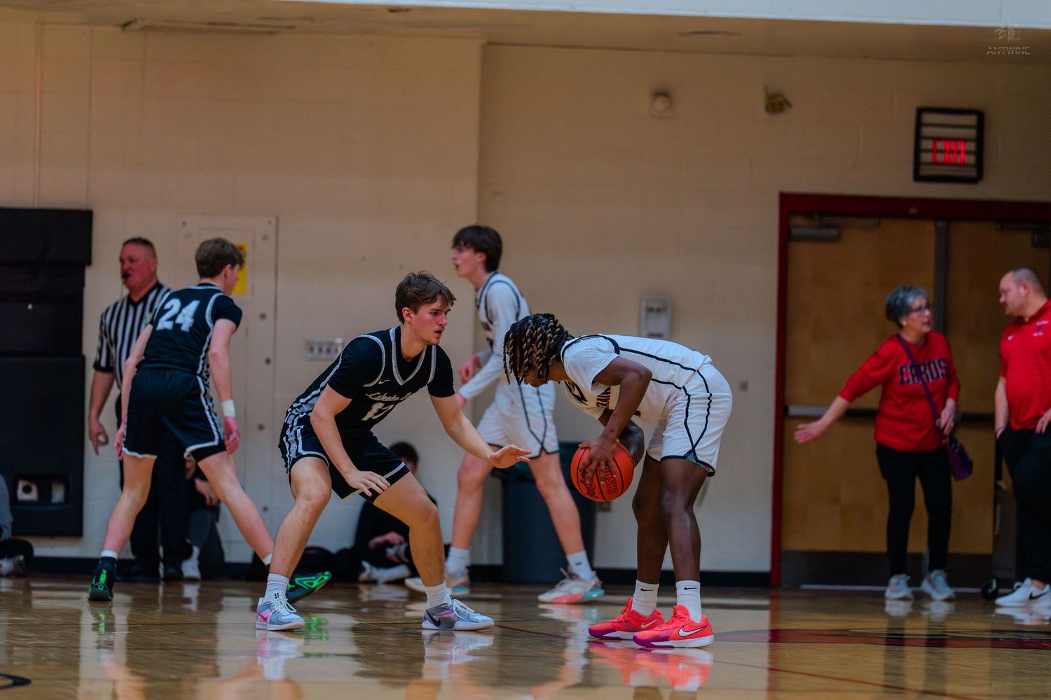 High school basketball players in defensive stance during game with referee and coach visible on sideline.