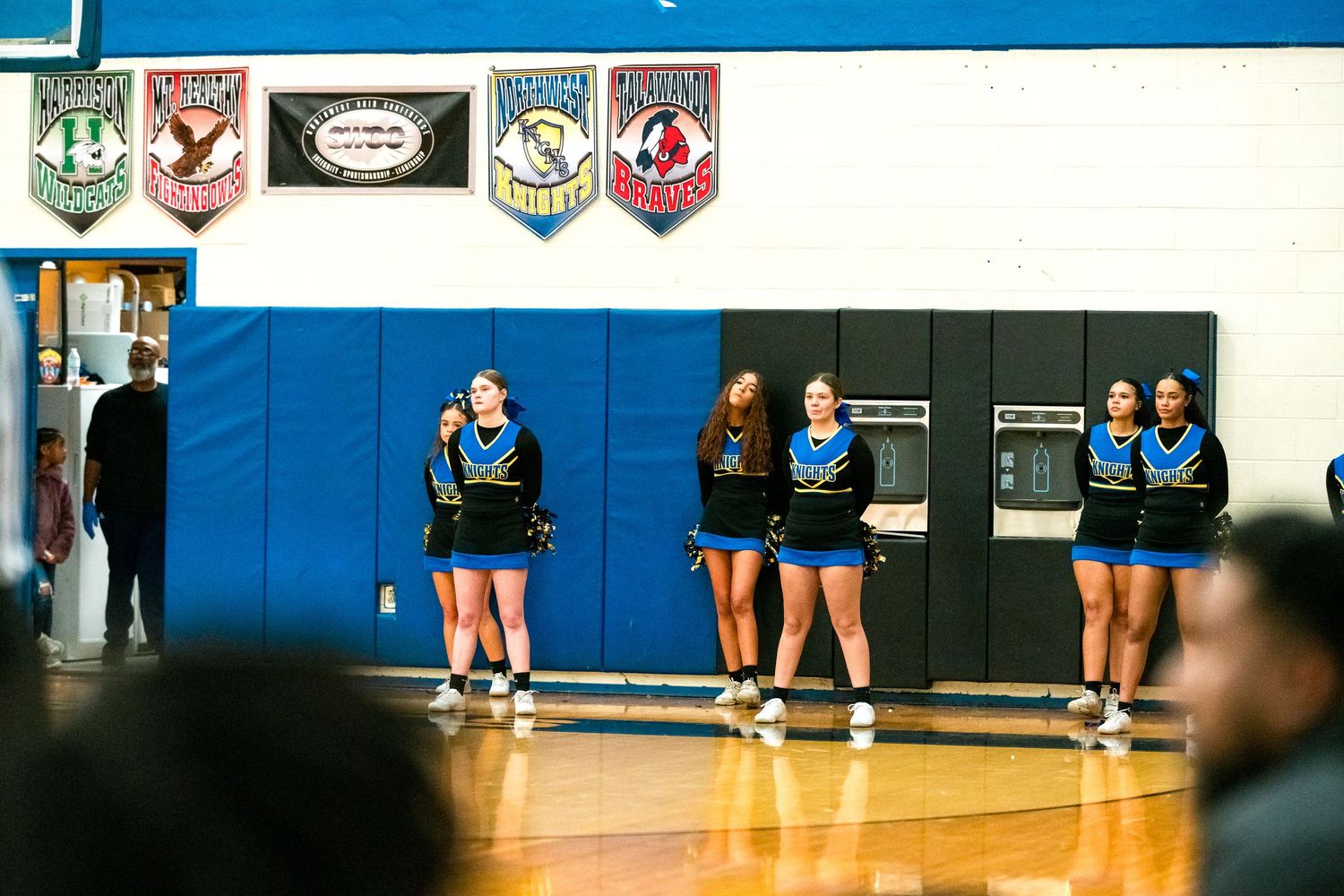Blue and black uniformed cheerleaders line up in a school gymnasium during a basketball game.