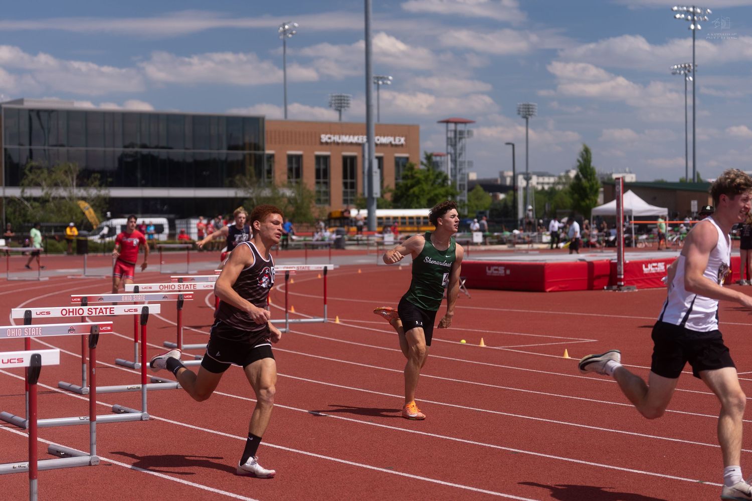 Three male athletes sprint past hurdles on the Ohio State University track during a championship hurdles race.
