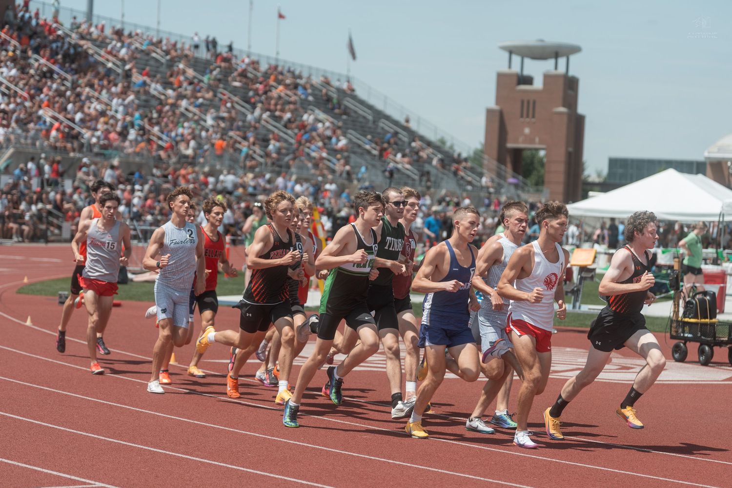 Large group of male athletes race together on a red track at a sunny high school track and field competition.