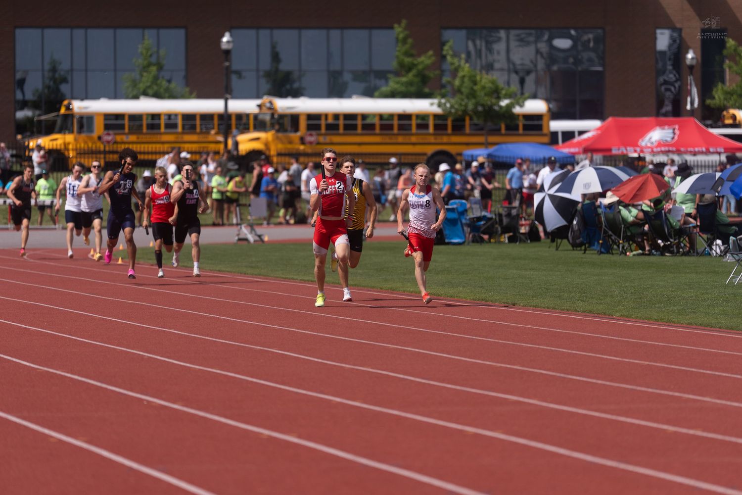 Competitive runners sprint on a red track during an outdoor high school track and field championship event.