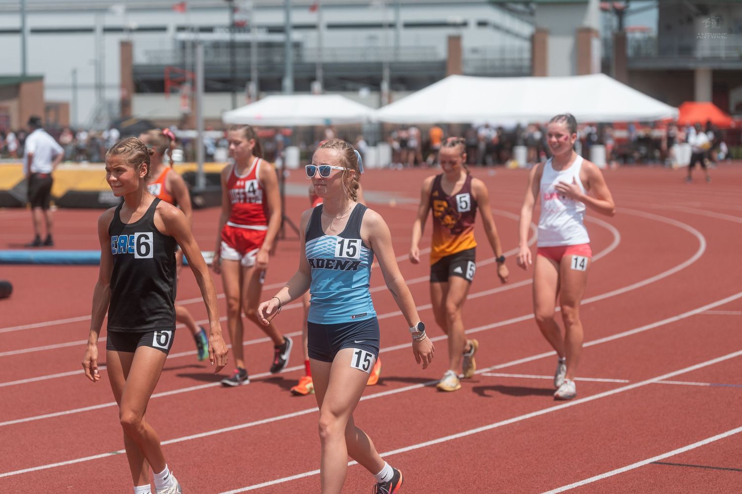 Female high school track athletes in numbered bibs competing in distance race on red outdoor track.