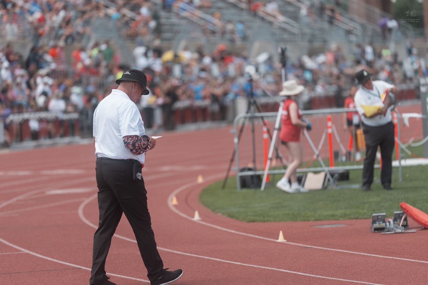 Track official reviews notes while walking along a red track curve at a packed outdoor stadium during a meet.