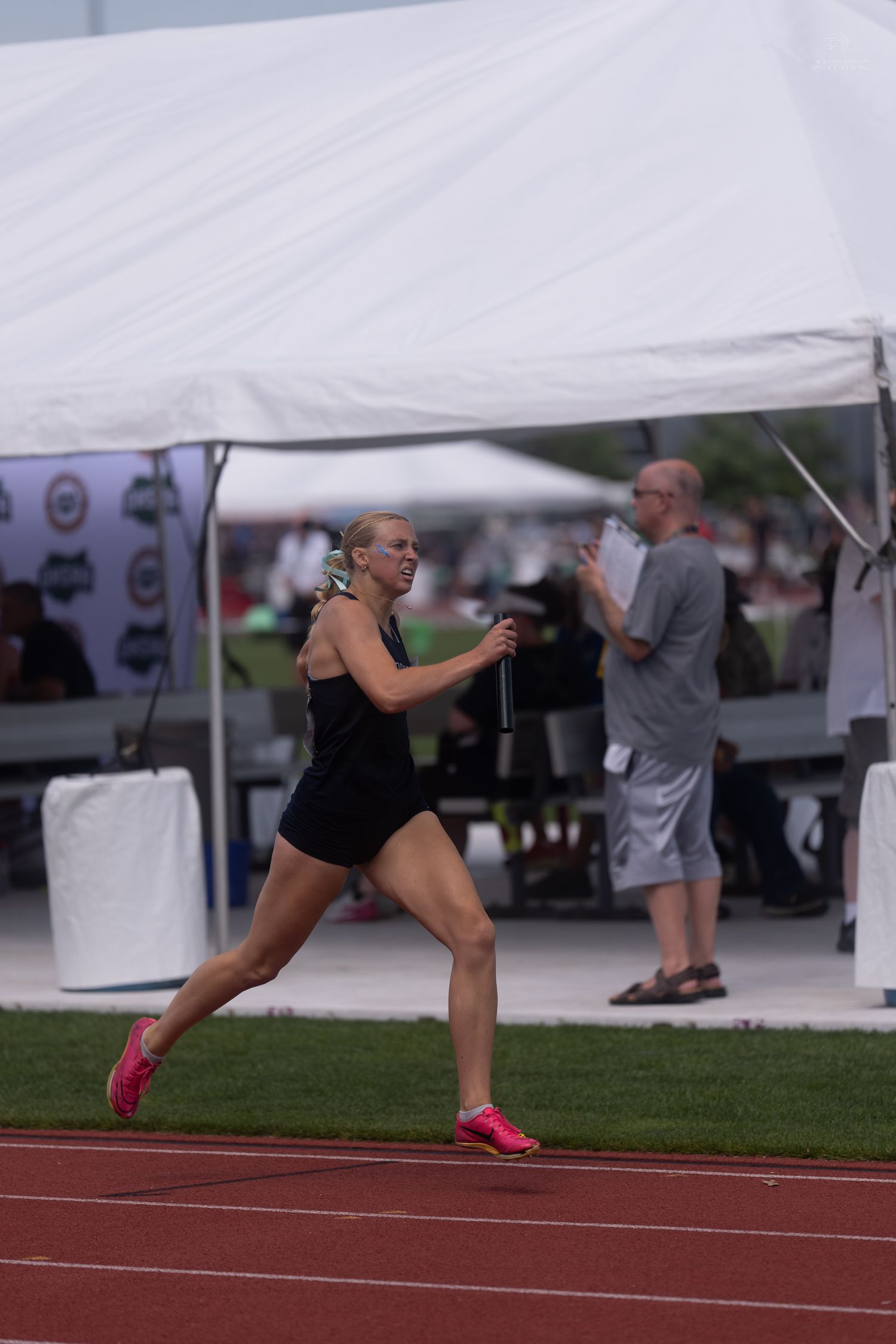Female relay runner in black uniform sprints on red track holding baton at outdoor track meet.
