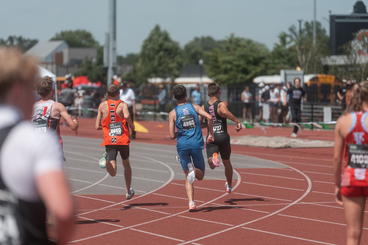 Male runners race down red track during outdoor track and field competition with crowd watching from sidelines.