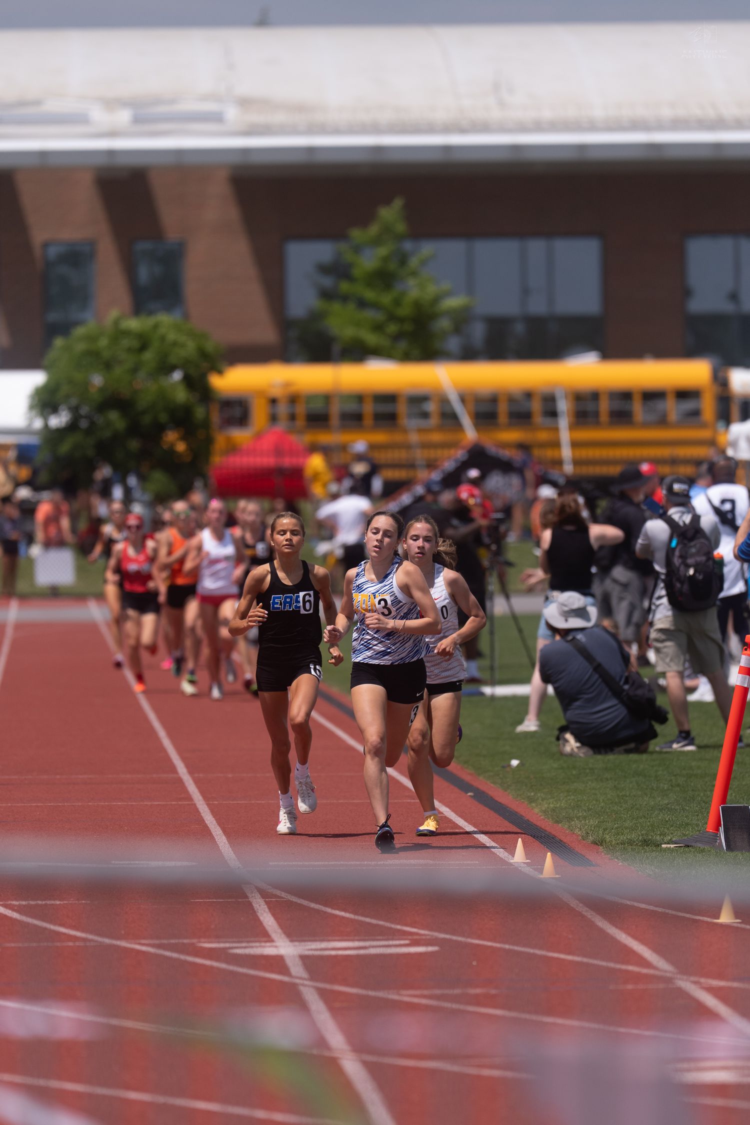 Female runners race on a red track during an outdoor high school track and field competition on a sunny day.