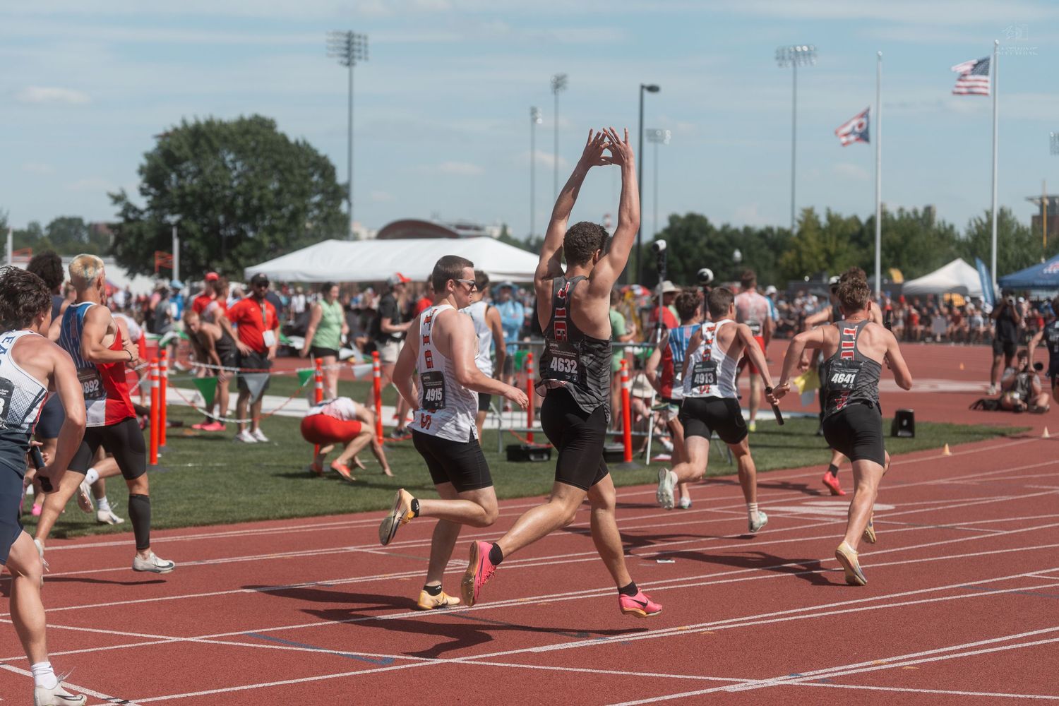 Runner raises baton overhead during relay exchange at competitive outdoor high school track and field event.