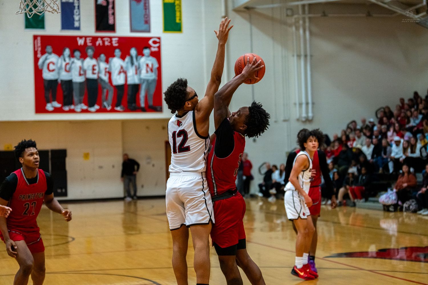 High school basketball player number 12 in white attempts shot while defender in red contests the ball.