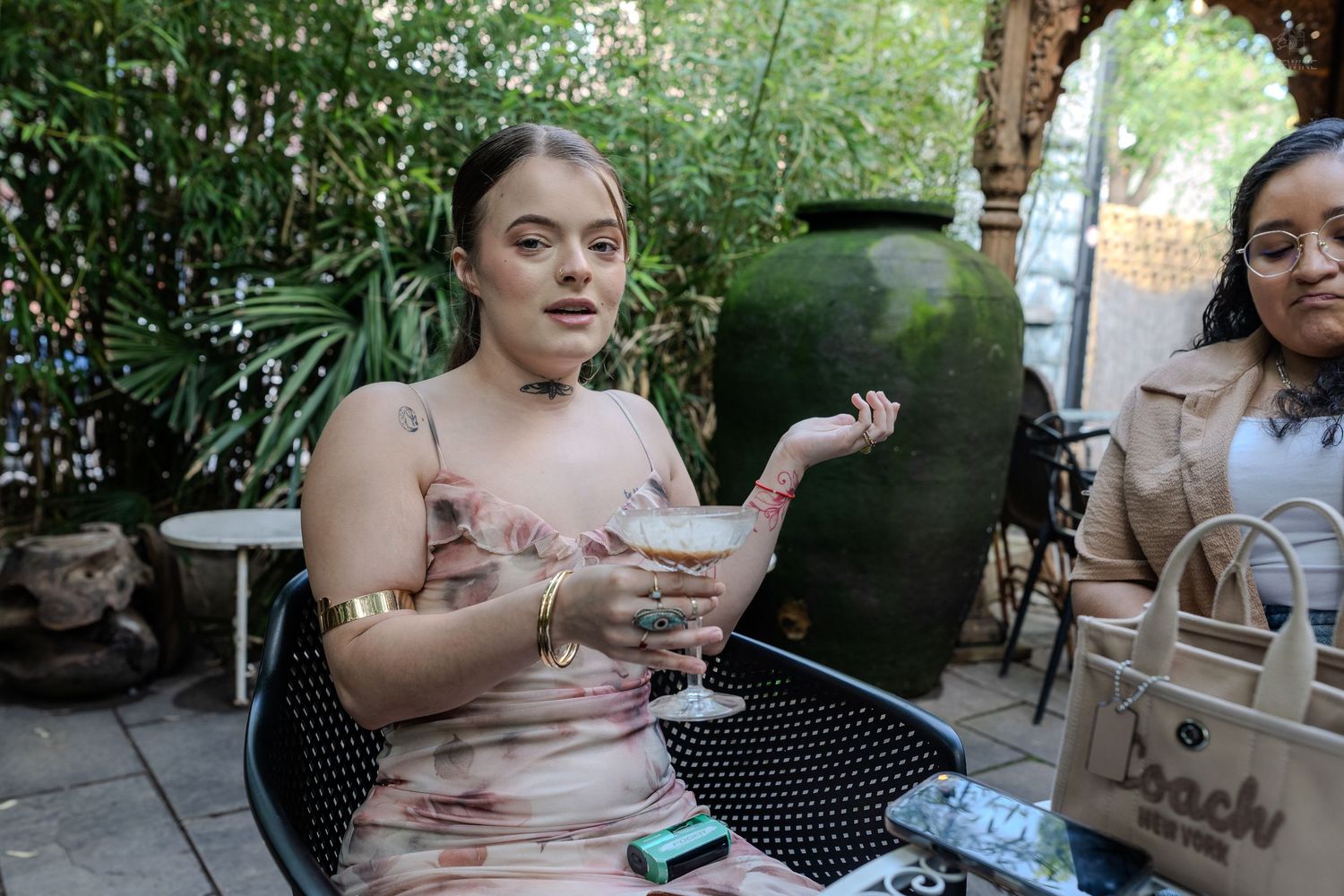 Woman in floral dress holding a cocktail glass at an outdoor patio with large decorative vase.