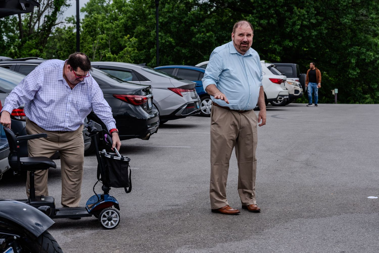 Two men in a parking lot, one handling a mobility scooter near parked cars.