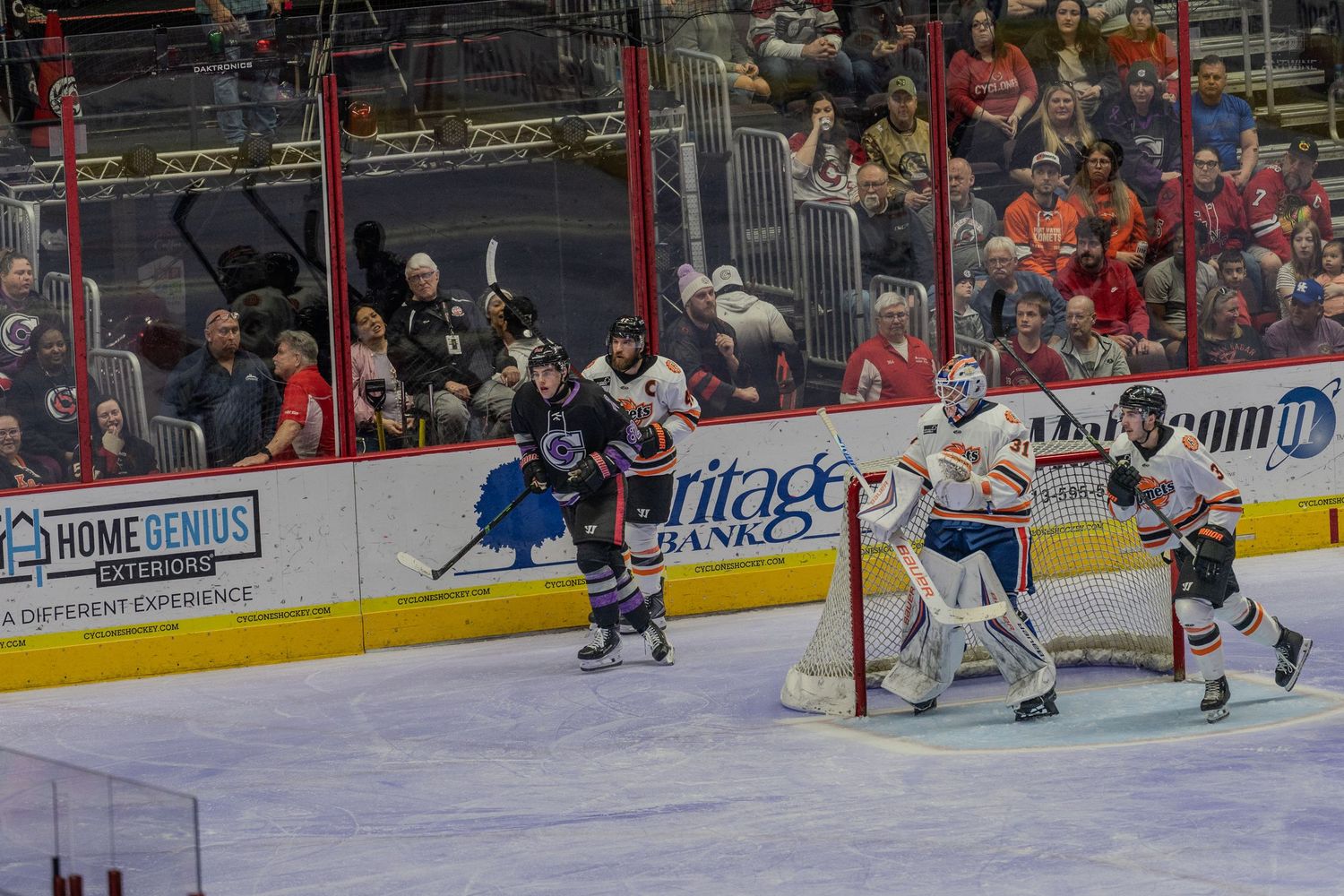 Ice hockey goalie in orange and white defends the net as opposing player approaches during a game.