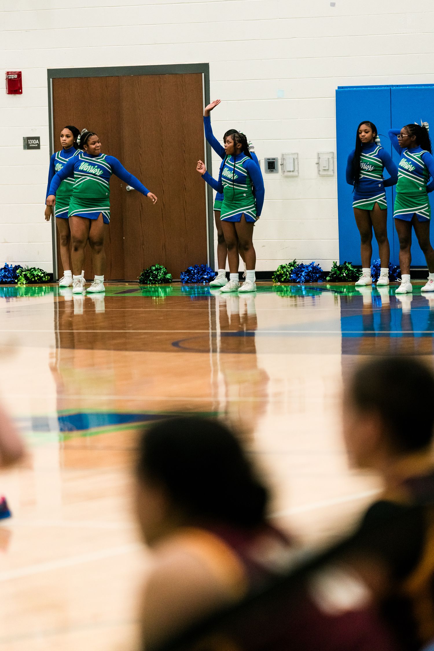 Cheerleaders in blue and green uniforms perform routines on a gymnasium floor during a high school basketball game.