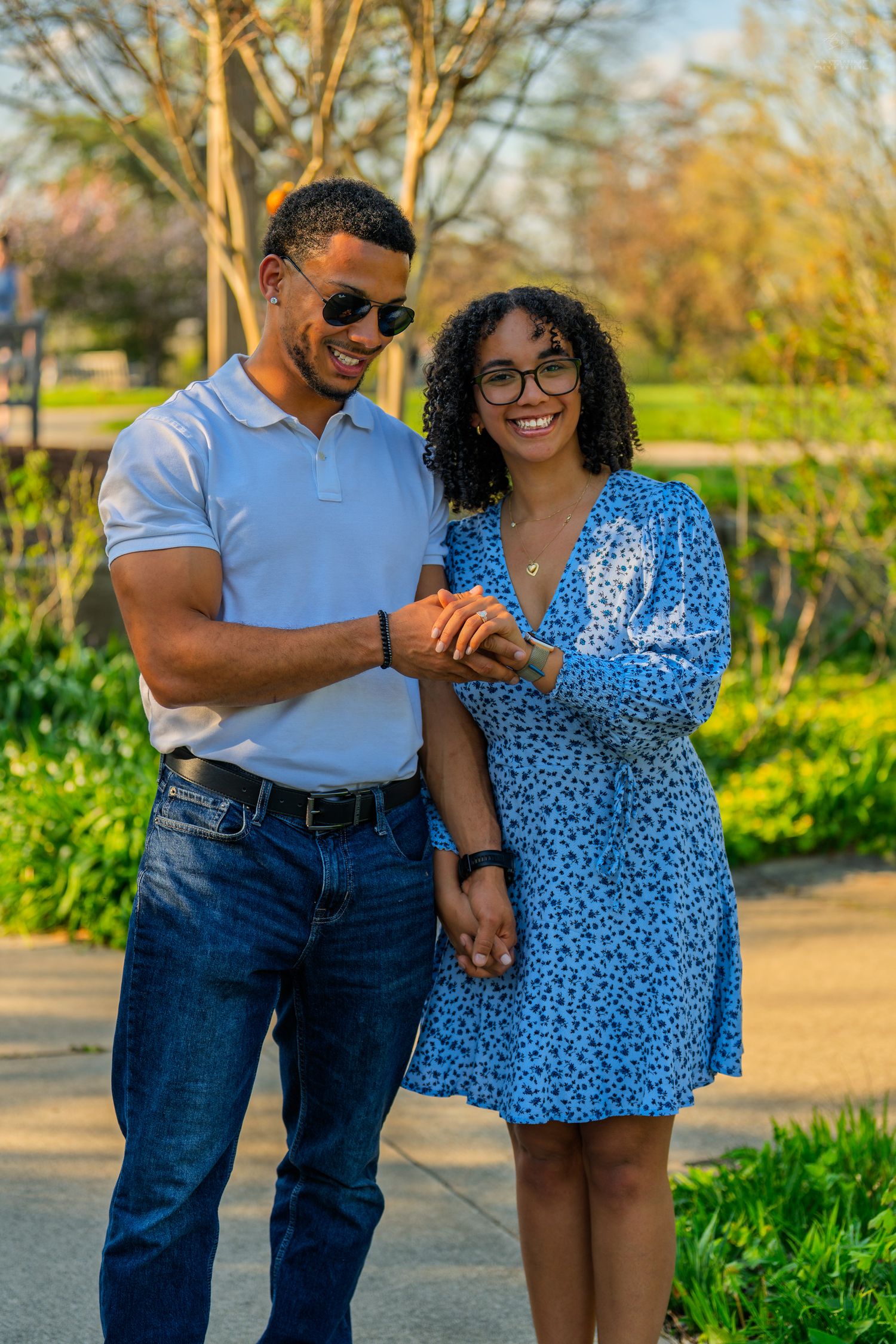 A smiling couple holds hands in a sunny park, the woman wearing a blue floral dress and glasses.