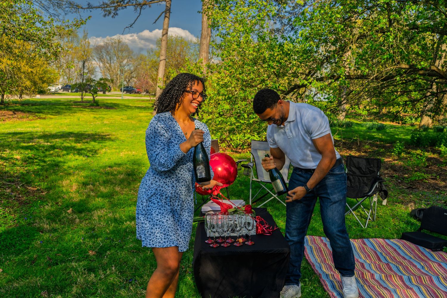 Couple at outdoor celebration table with red balloons, champagne bottle and glasses in sunny park.