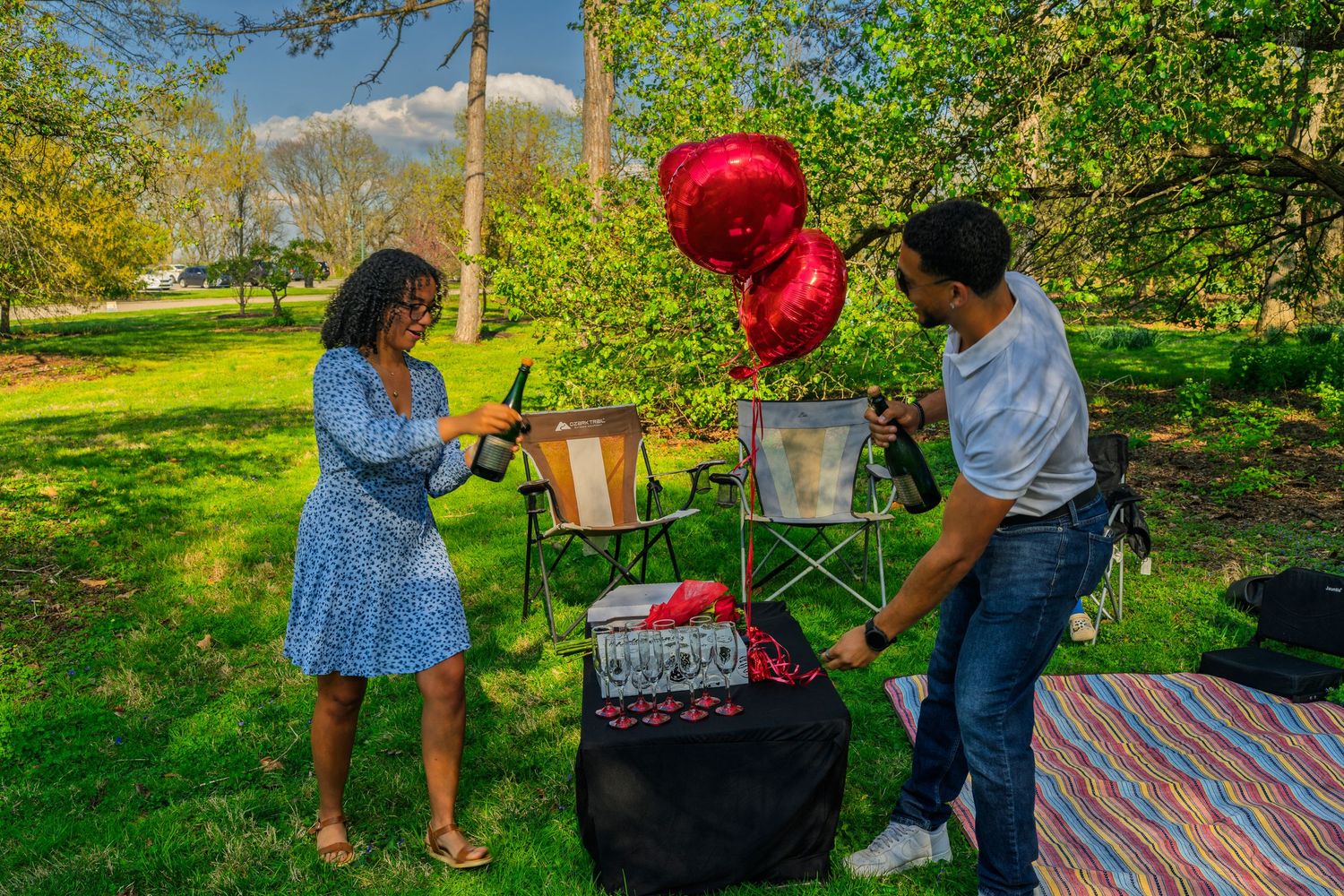 Couple enjoying a romantic outdoor picnic with red heart balloons and champagne bottles in a lush green park.