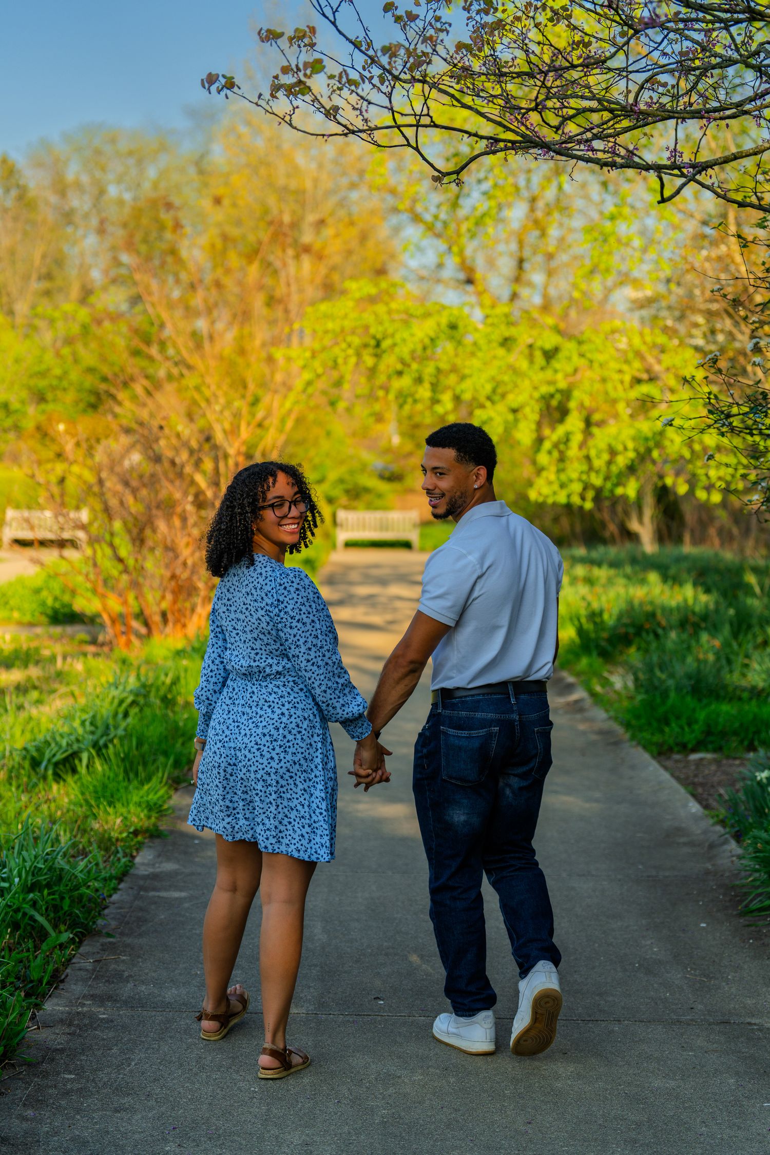 Couple holding hands and looking at each other while walking on a sunlit park pathway.