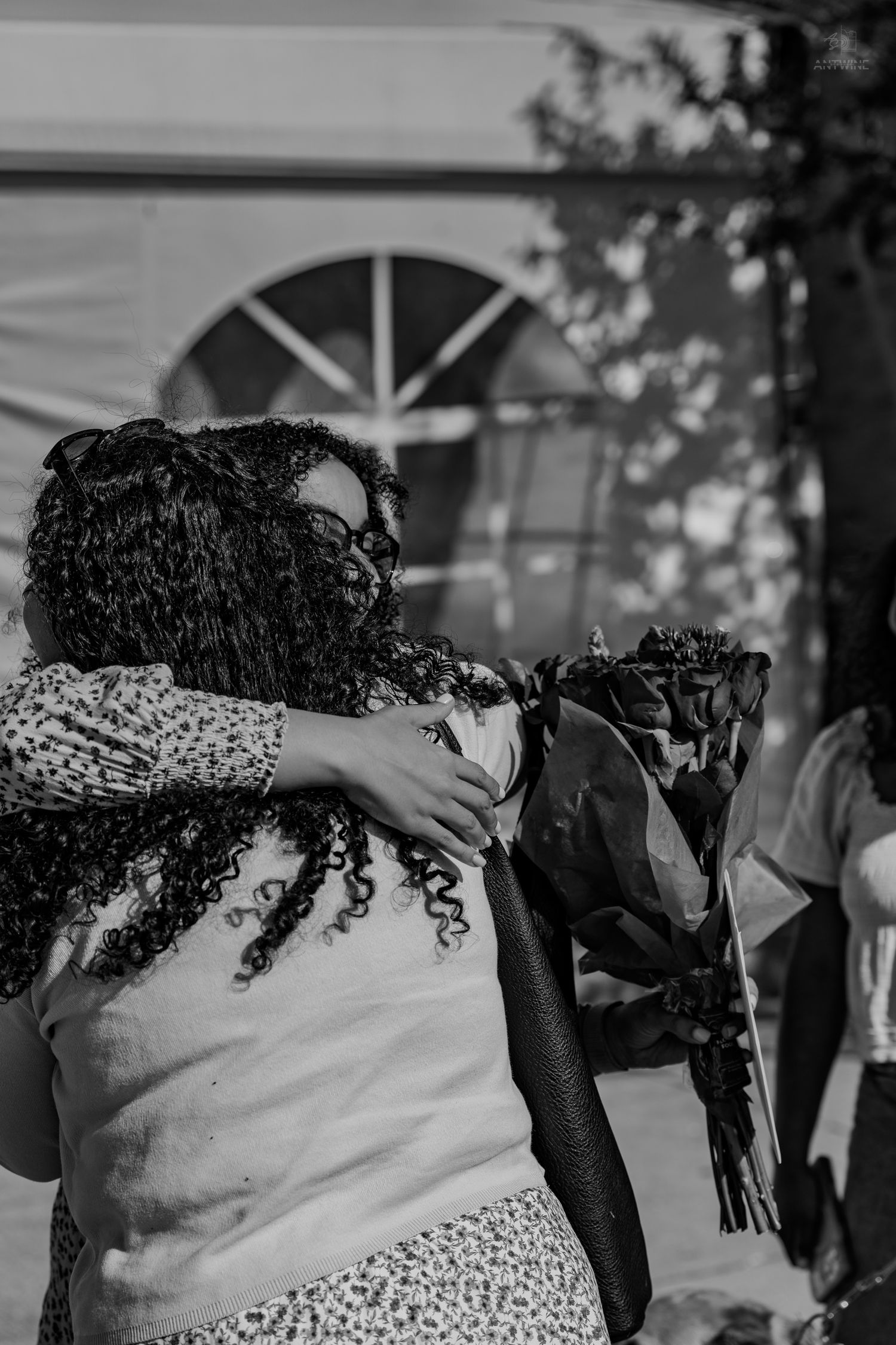 Two women share a warm embrace while holding a bouquet of flowers in black and white.
