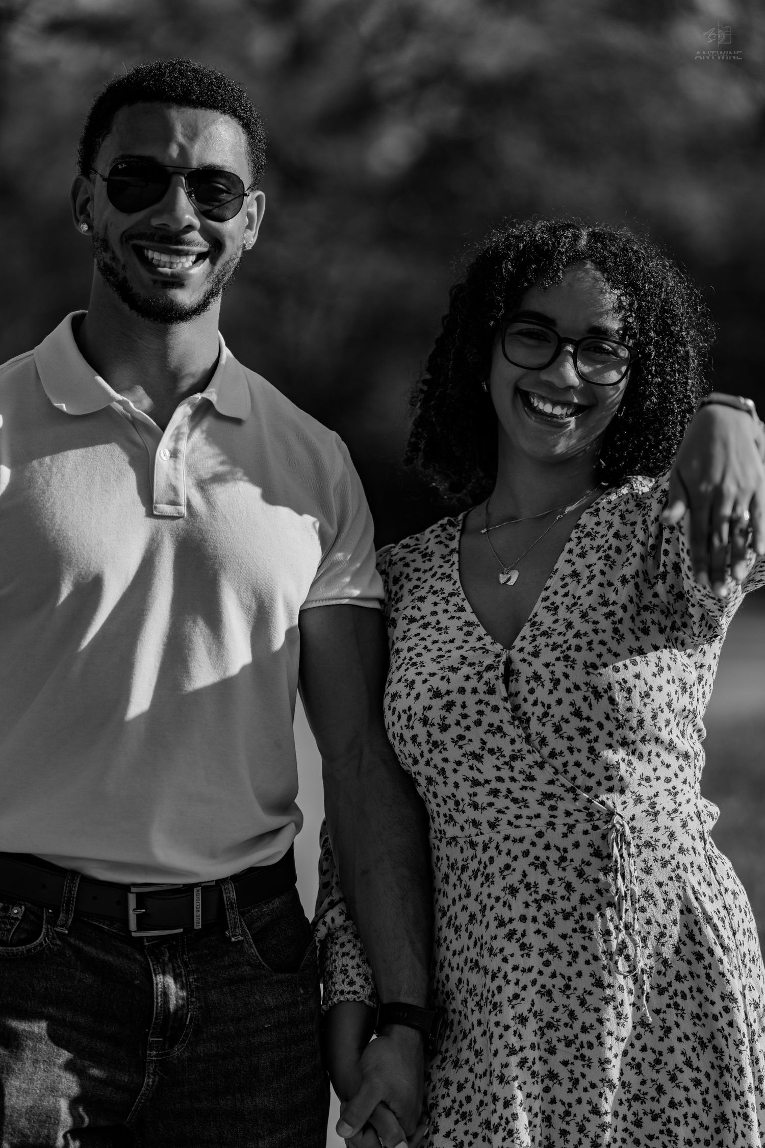 Black and white close-up of a smiling couple holding hands, man in sunglasses and woman in floral dress.