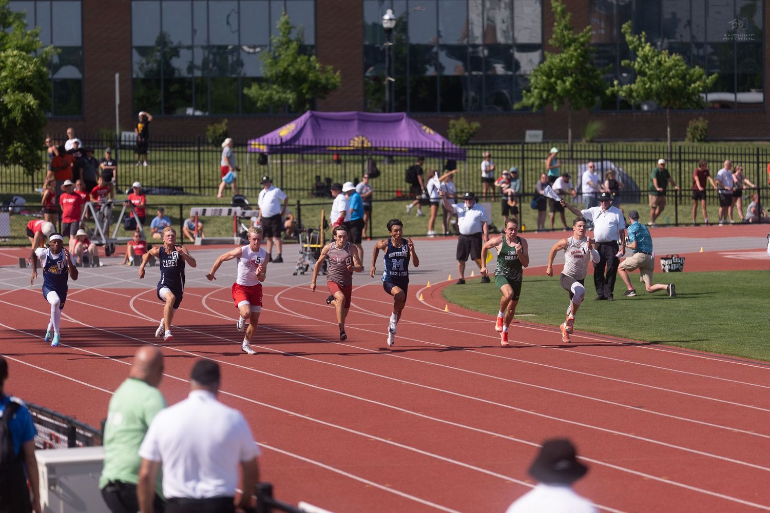 Young male runners compete in a sprint race on a red outdoor track during a sunny track and field meet.