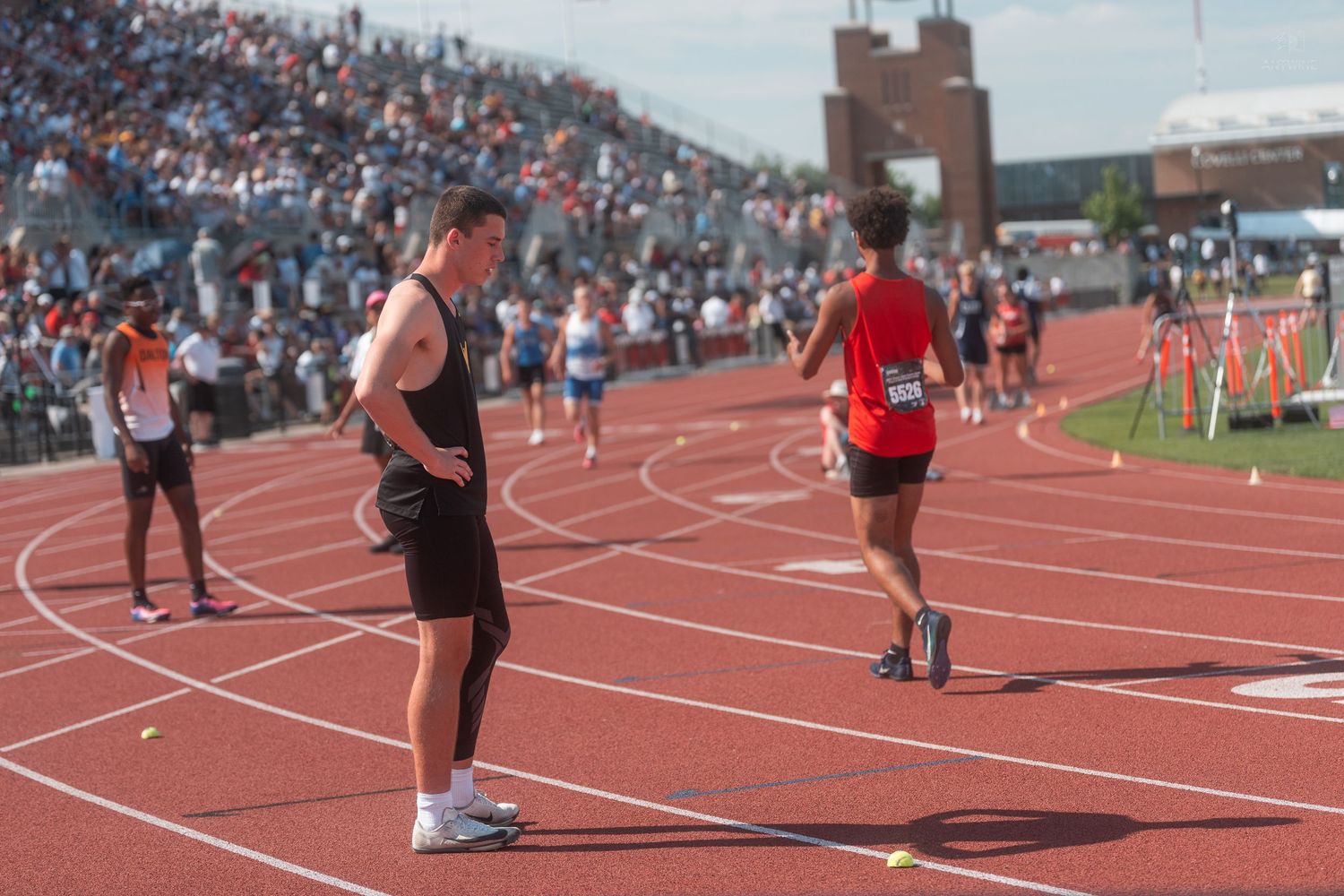 Coach in black shorts observes runners on red track during a large outdoor track and field meet.