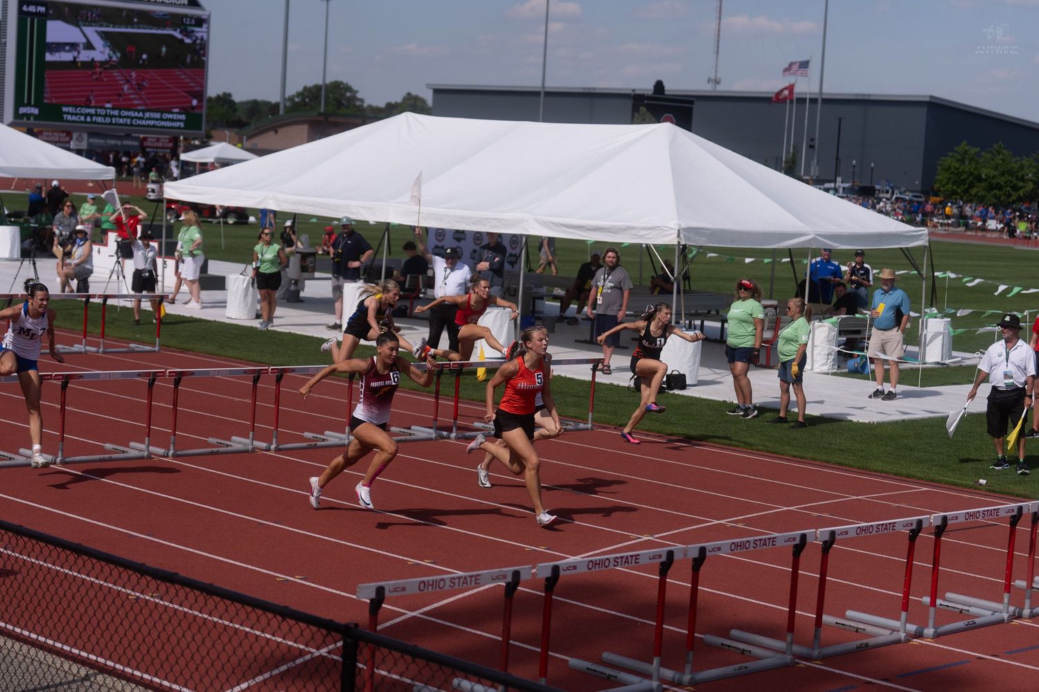 Female athletes competing in hurdles race on red track at outdoor high school track and field championship meet.